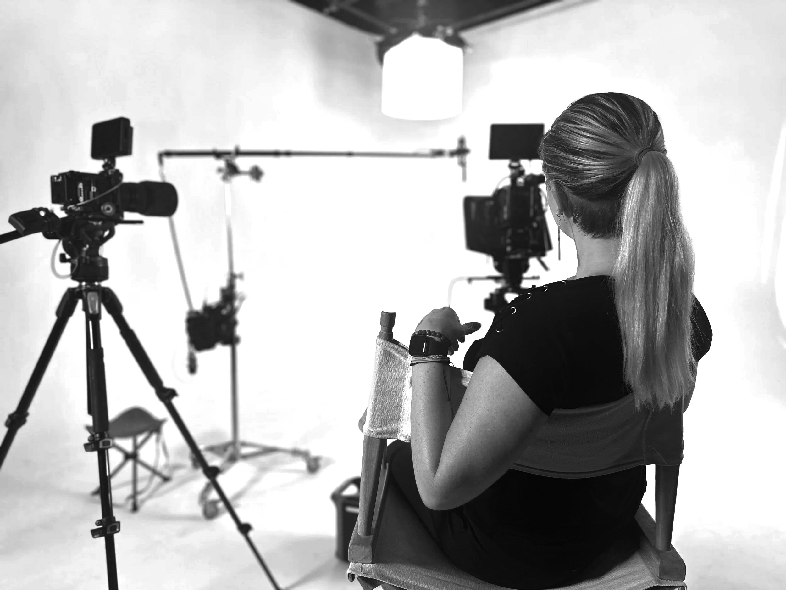 Black and white photo of a woman with long hair tied in a ponytail sitting on a chair in a photography studio, with professional cameras and lighting equipment set up around her.