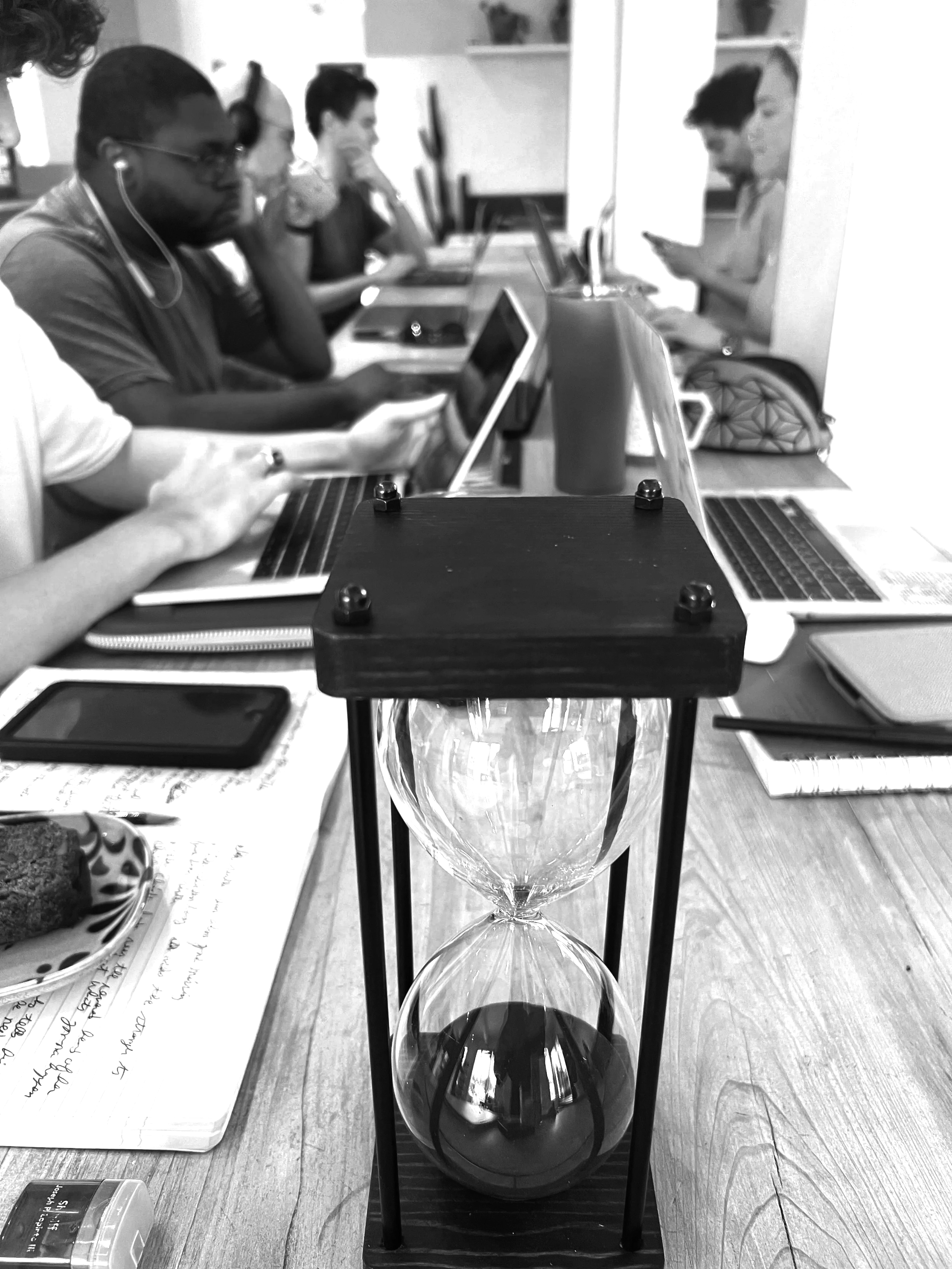 A lab setup for a science experiment, featuring an hourglass, a small electronic device, and a glass container, with people working and studying at a table in the background.