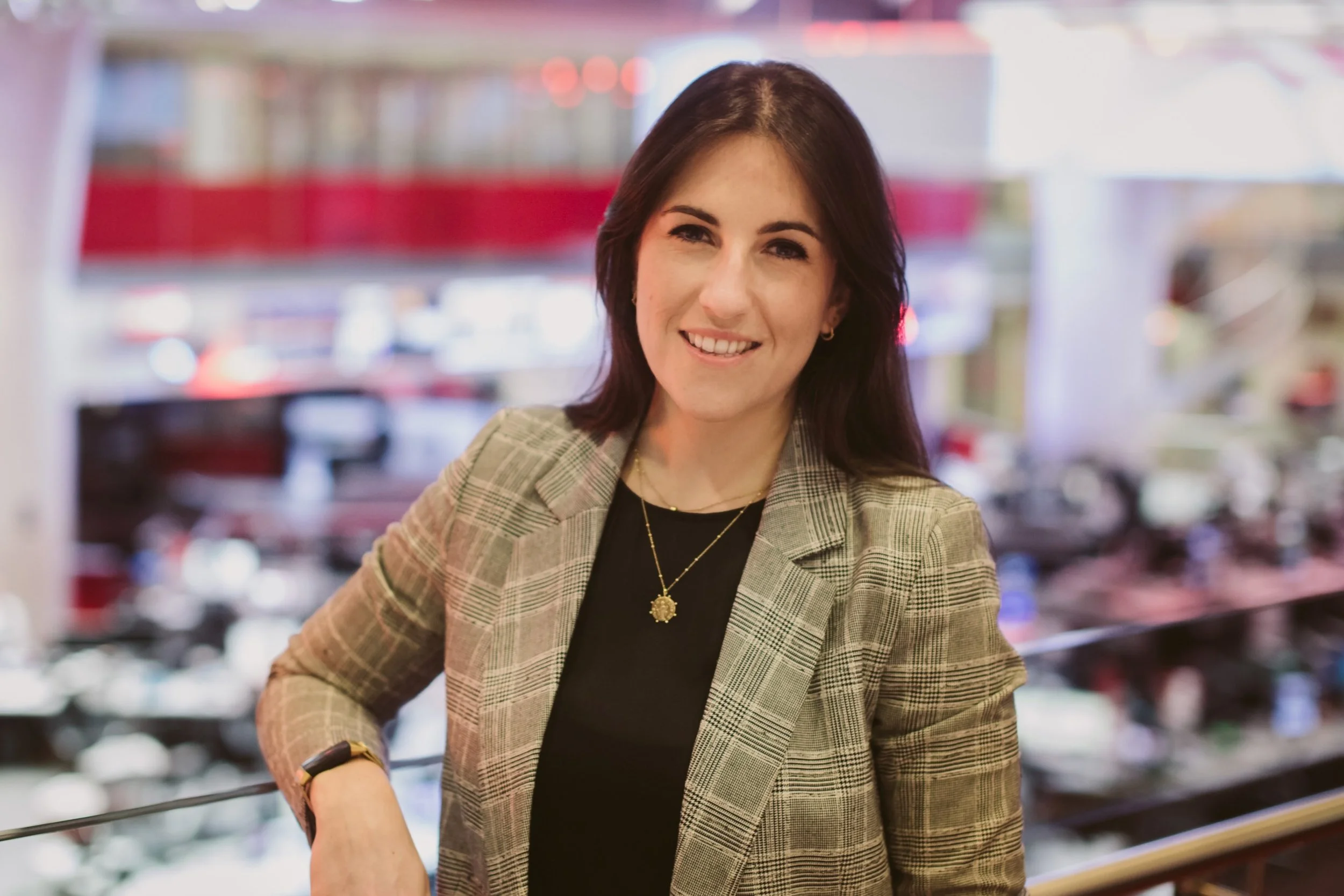 A woman with long dark hair smiling, wearing a checked blazer over a black top with a gold necklace, in a BBC Newsroom with desks and screens in the background.