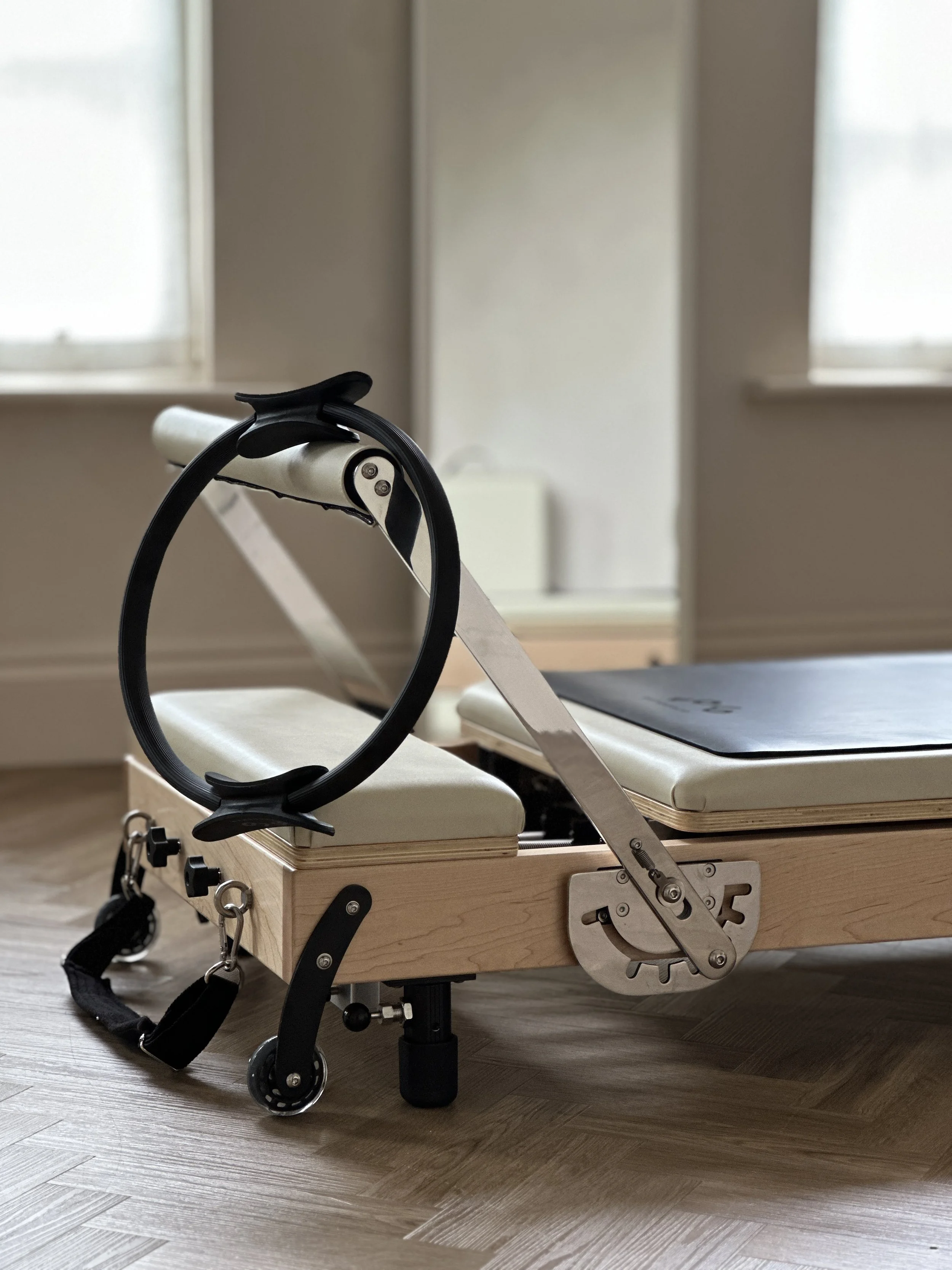 The corner of a beige leather Reformer Pilates machine with in a room with windows, a long mirror and hardwood flooring.