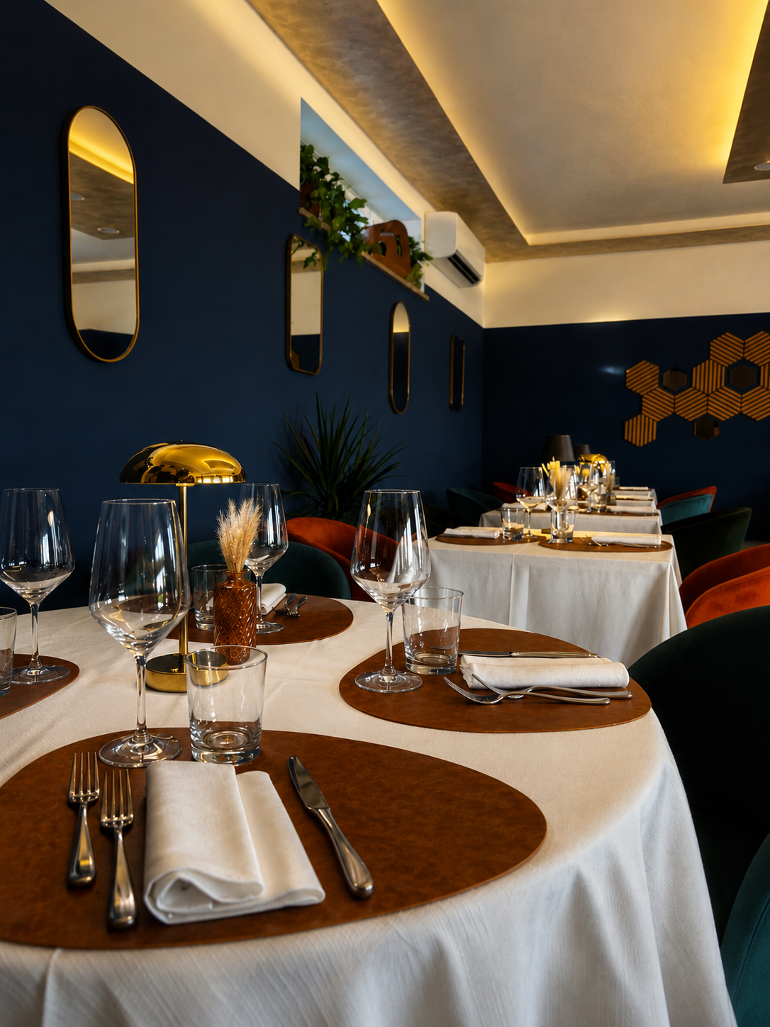 Tavoli apparecchiati con bicchieri, posate, tovaglioli e lampade in un ristorante elegante con pareti blu e specchi.