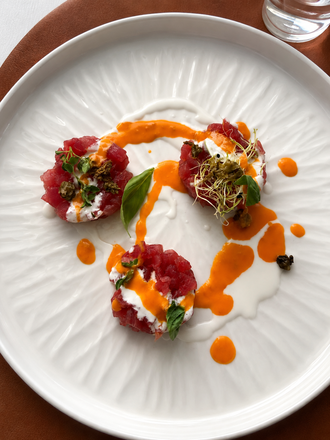 Piatti di tartare di tonno con salsa di panna, olio di peperoncino, microgreens, basilica, e pepe di capra, serviti su un piatto bianco.