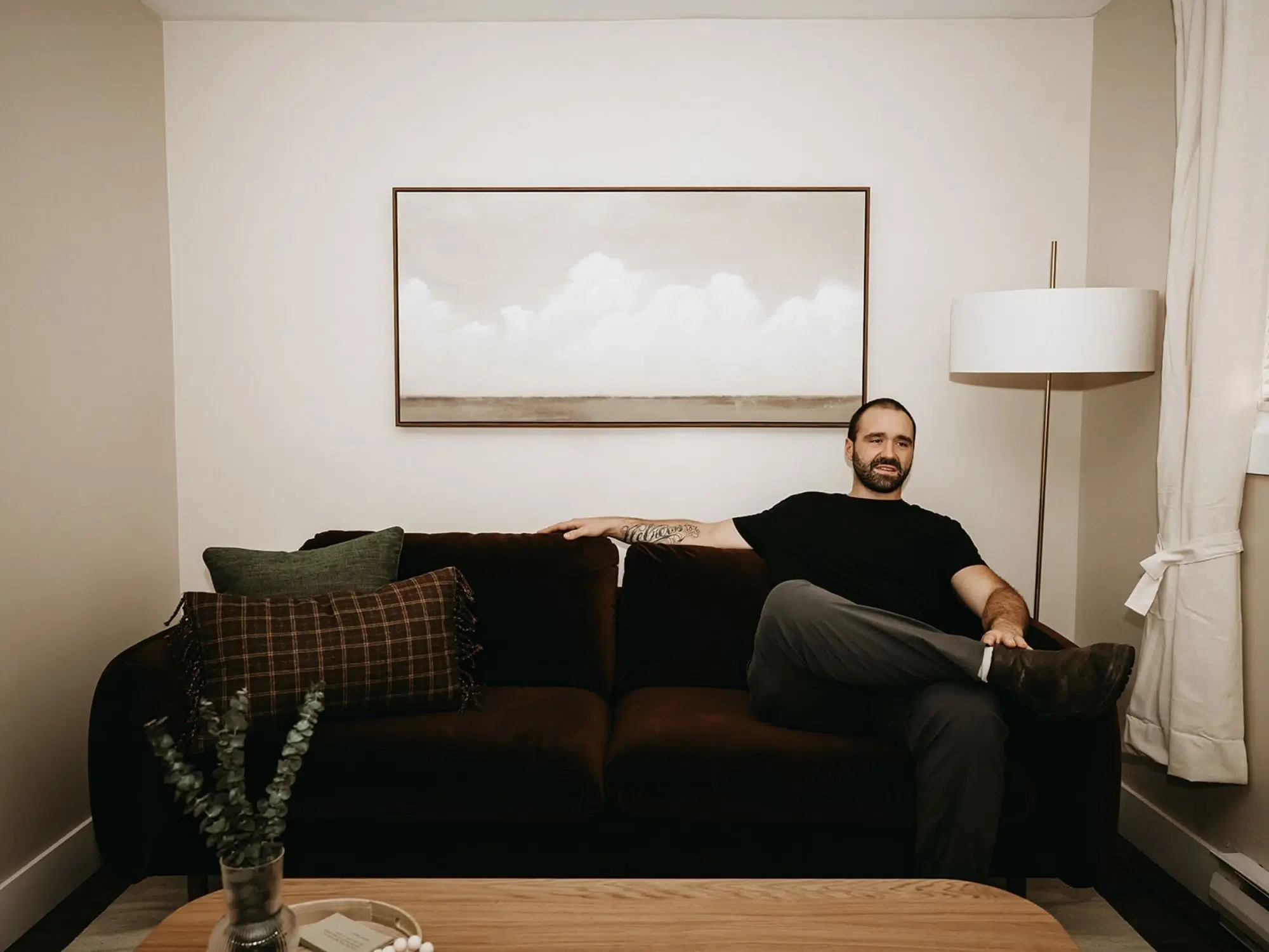 Man with beard and tattoos reclining on a dark sofa in a living room with white walls, a landscape painting, and a tall floor lamp next to a window with curtains.