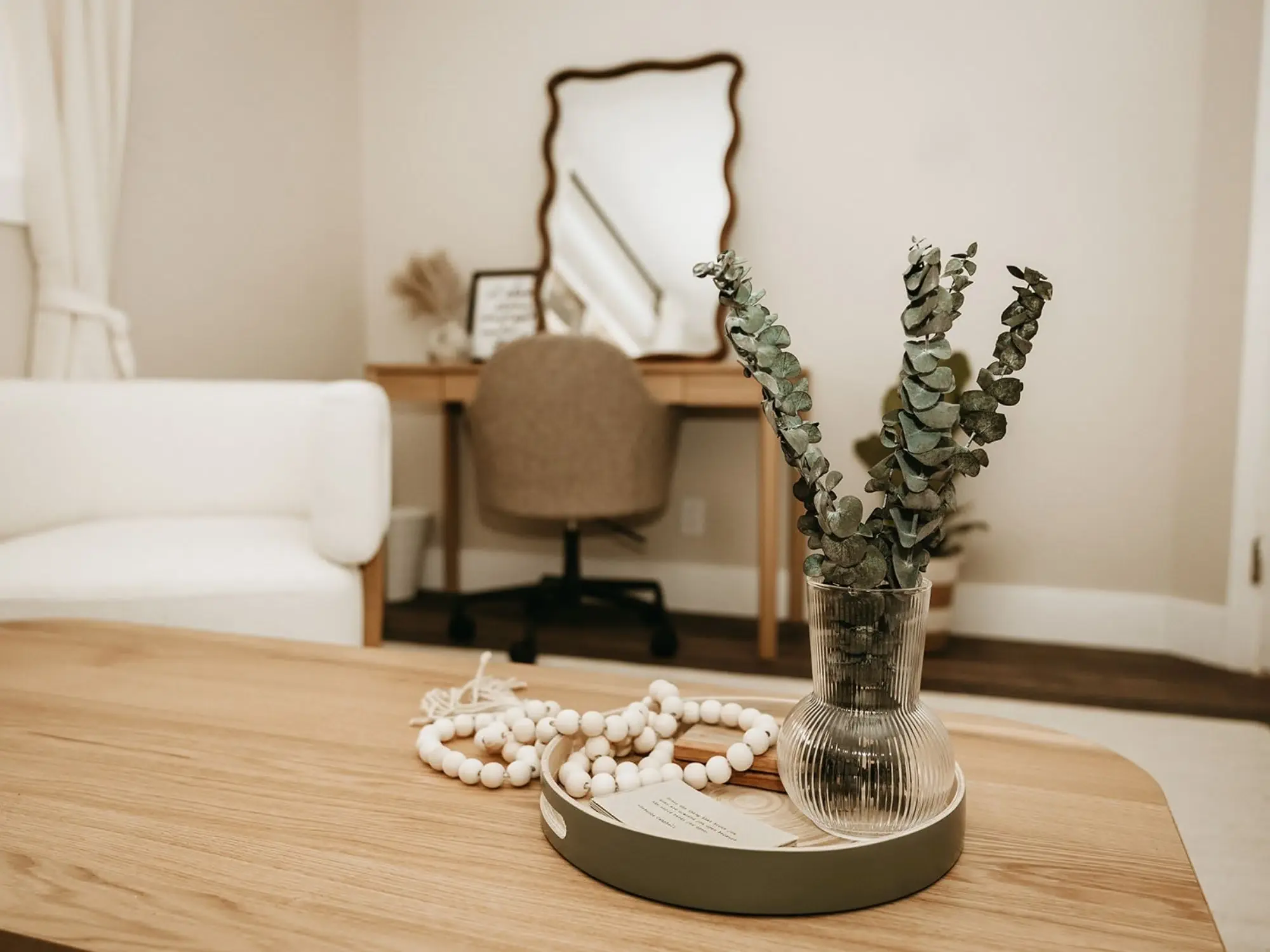 Decorative arrangement on a wooden table includes a glass vase with eucalyptus, a string of white beads, and a small white card on a tray, with a cozy living space background featuring a white sofa, a desk with a mirror, and a beige chair.