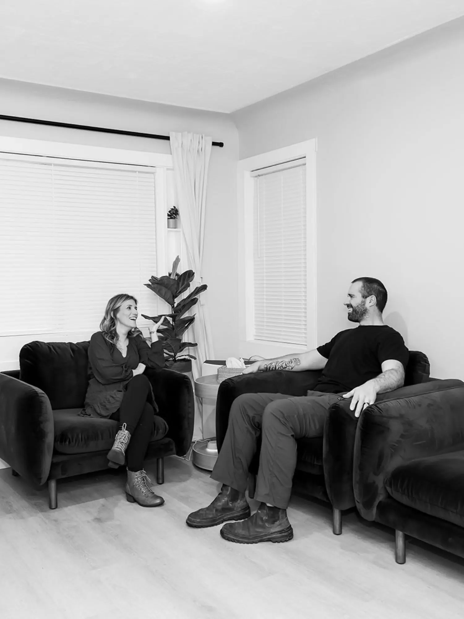 A man and woman sitting on separate couches facing each other and smiling. The woman has shoulder-length hair, is wearing a long-sleeve shirt, and lace-up boots. The man has a beard, tattoos on his arm, and is wearing a black T-shirt and cargo pants. There is a table with a tissue box and some electronic device between them in a living room with a window, blinds, curtains, and a potted plant.