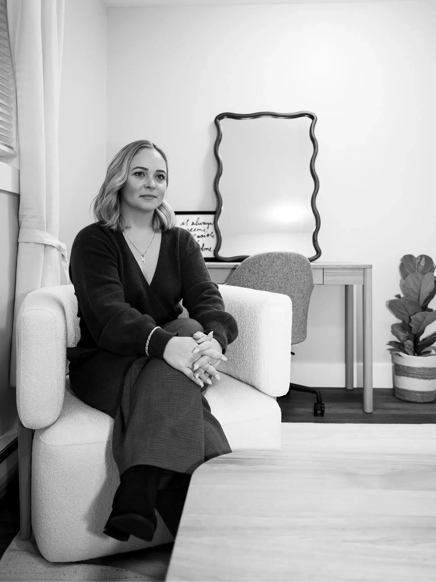 A woman sitting on a white sofa in a modern, minimalist office or living space, with a desk, chair, mirror, and potted plant in the background, all in black and white.