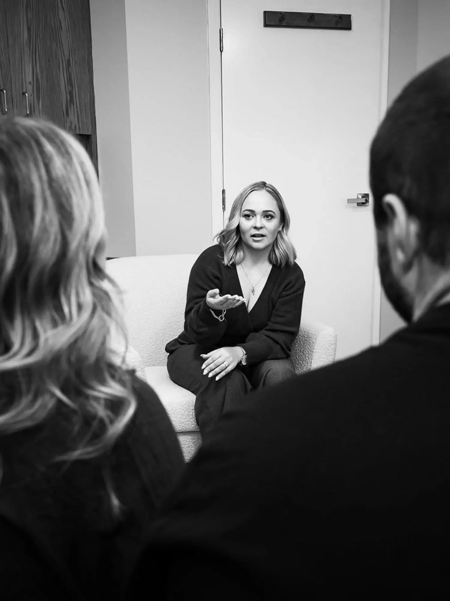 A woman is sitting on a sofa, gesturing with her hand, engaged in conversation with two people whose backs are to the camera in a room with plain walls and a closed door.