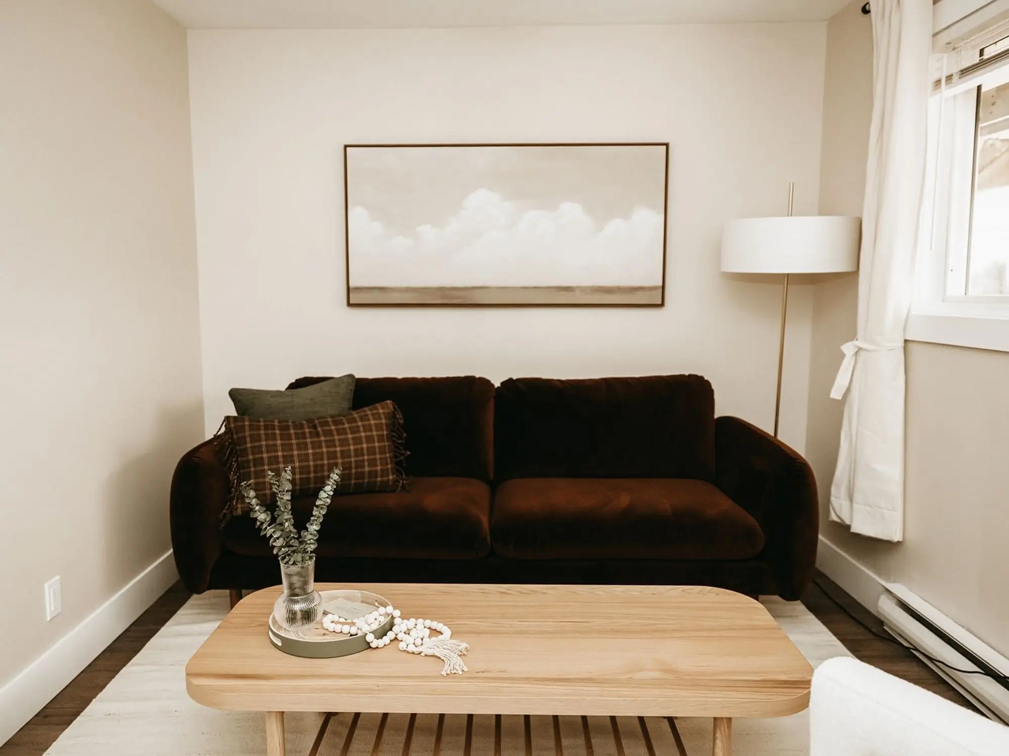 Living room with a brown sofa, wall art, a floor lamp, a window with white curtains, and a wooden coffee table with decorative items.