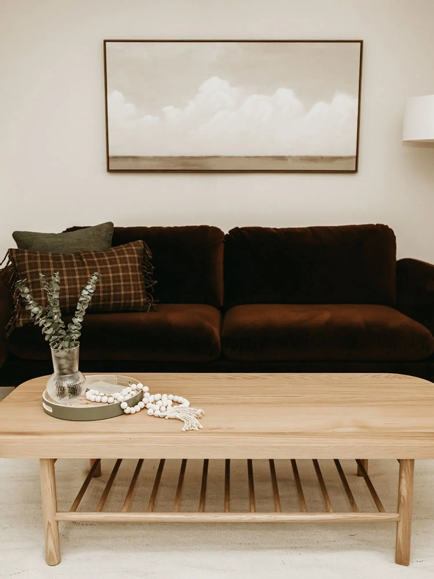 A living room scene with a dark brown sofa, a large framed landscape painting of clouds above a landscape, a wooden coffee table, a vase with dried flowers, and a string of white beads.