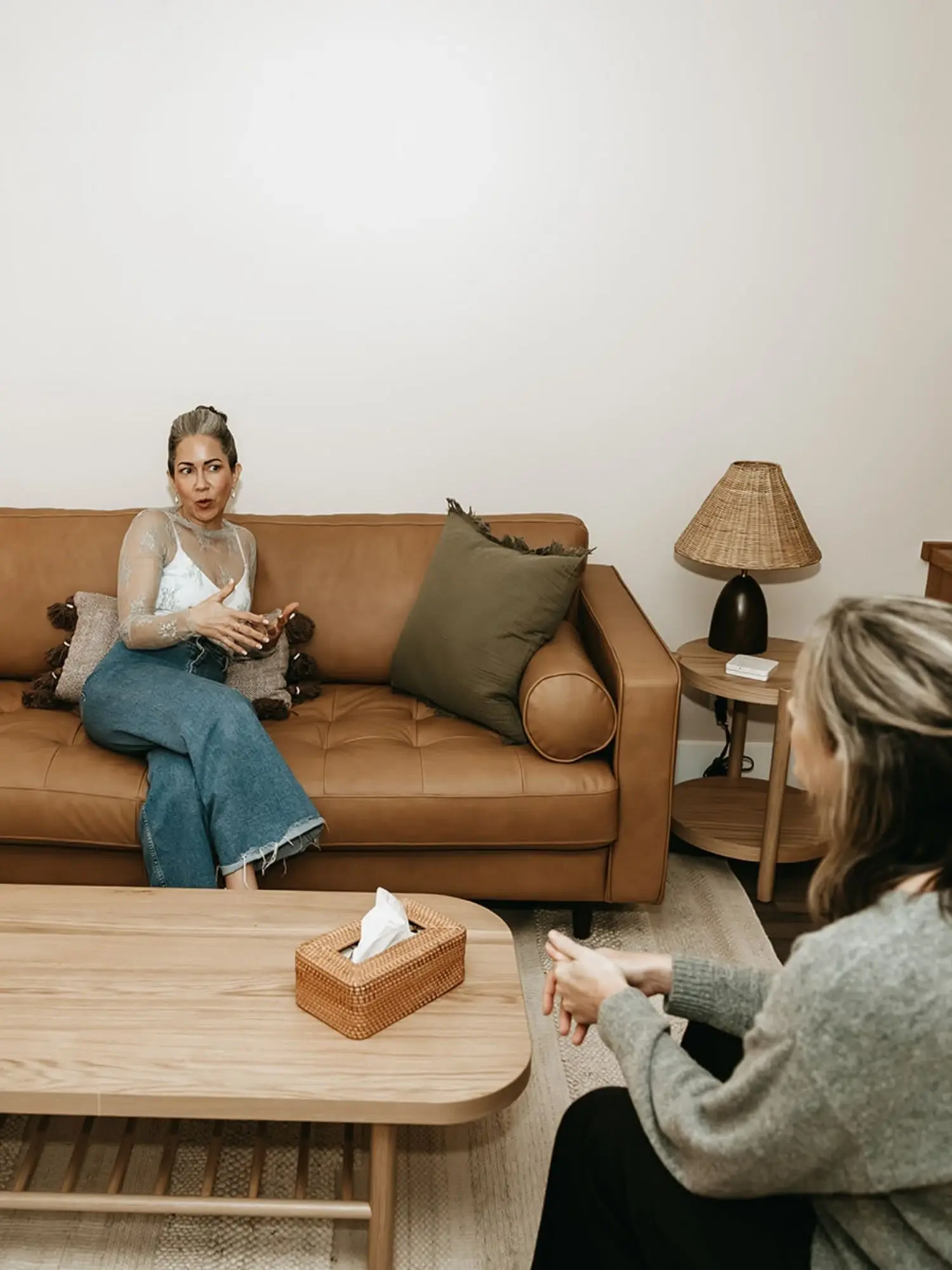 Two women having a conversation in a living room; one is sitting on a tan leather sofa, and the other is seated on a chair. There is a wooden coffee table with a tissue box, a side table with a lamp, and a plain wall in the background.