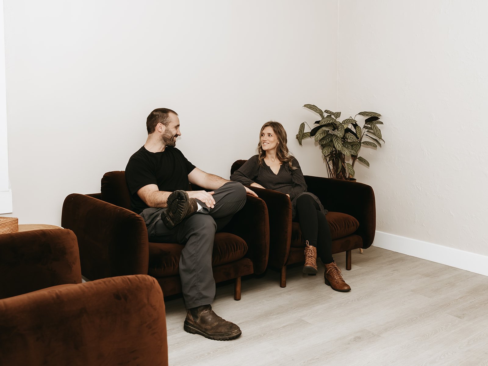 A man and a woman sitting on a brown sofa, smiling and engaging in conversation in a minimalistic room with white walls and a potted plant.
