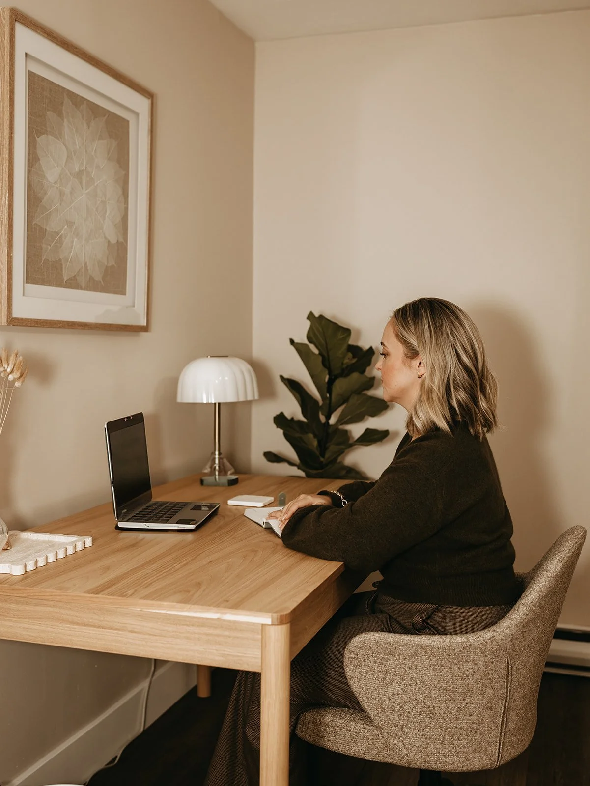 Woman sitting at a wooden desk with a laptop, notebook, and pen, in a cozy home office with a framed artwork, a table lamp, and a large plant in the background.