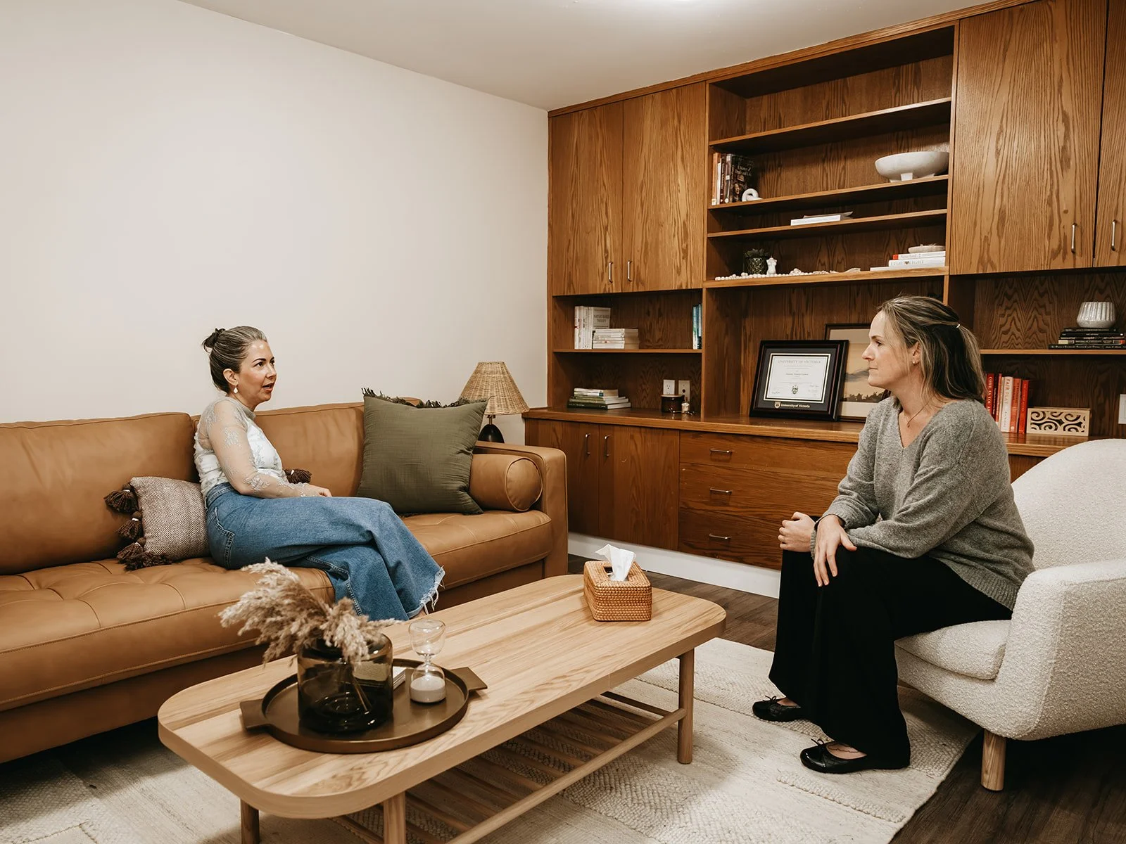 Two women having a conversation in a cozy living room. One is sitting on a tan leather sofa, the other on a white armchair. The room features wood furniture and shelves with books and decor.