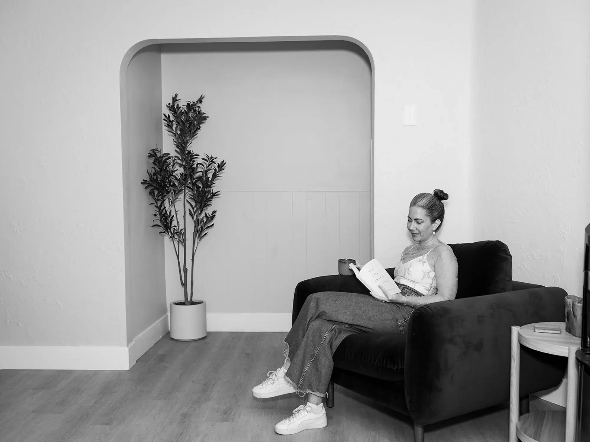 A woman sitting on a black sofa, reading a book and holding a cup in a cozy living room, with a potted plant and side table nearby.