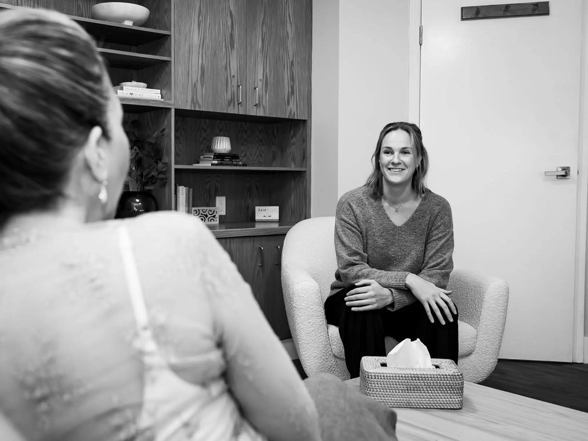 A woman with shoulder-length hair sitting in a plush armchair, smiling during a conversation with another woman whose back is to the camera, in an office setting with a wooden bookshelf in the background.
