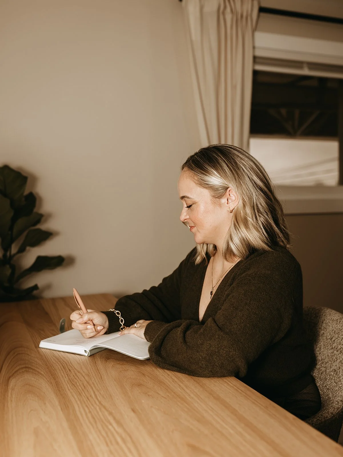 A woman writing in a notebook at a wooden table, with a plant and a window with curtains in the background.