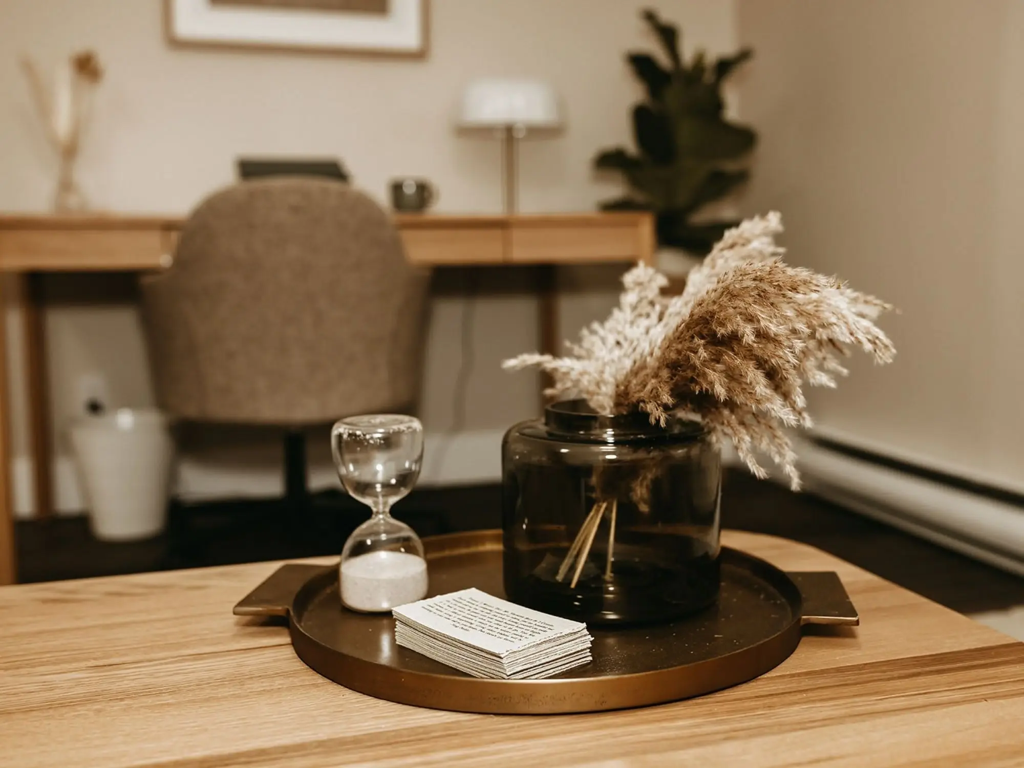 Decorative table setting with a black vase holding beige pampas grass, a glass hourglass, and a small stack of paper notes on a round tray, on a wooden table.