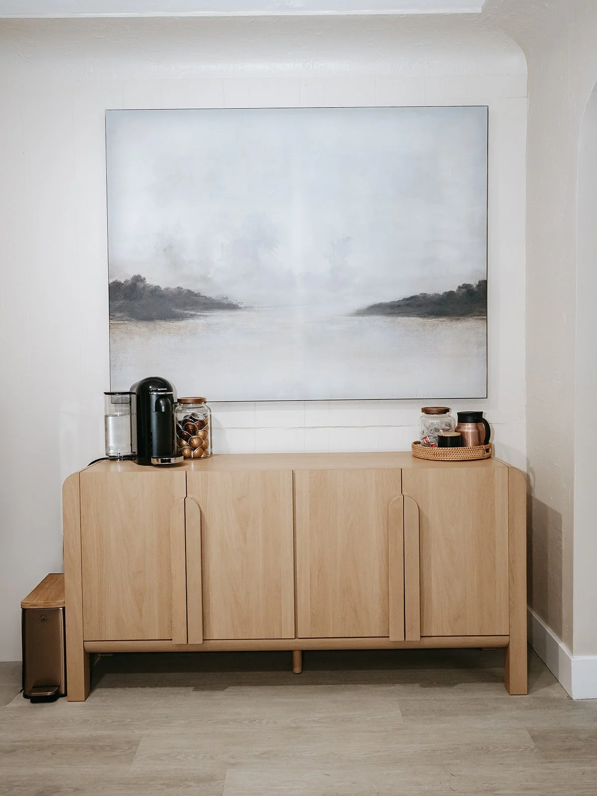 A light wood sideboard against a white wall with a large abstract landscape painting above it. On the left side of the sideboard, there is a black coffee machine, a jar with coffee capsules, and an empty glass. On the right side, there is a tray with jars, a small black and copper container, and a copper mug. A small trash bin is partially visible on the left side of the sideboard.