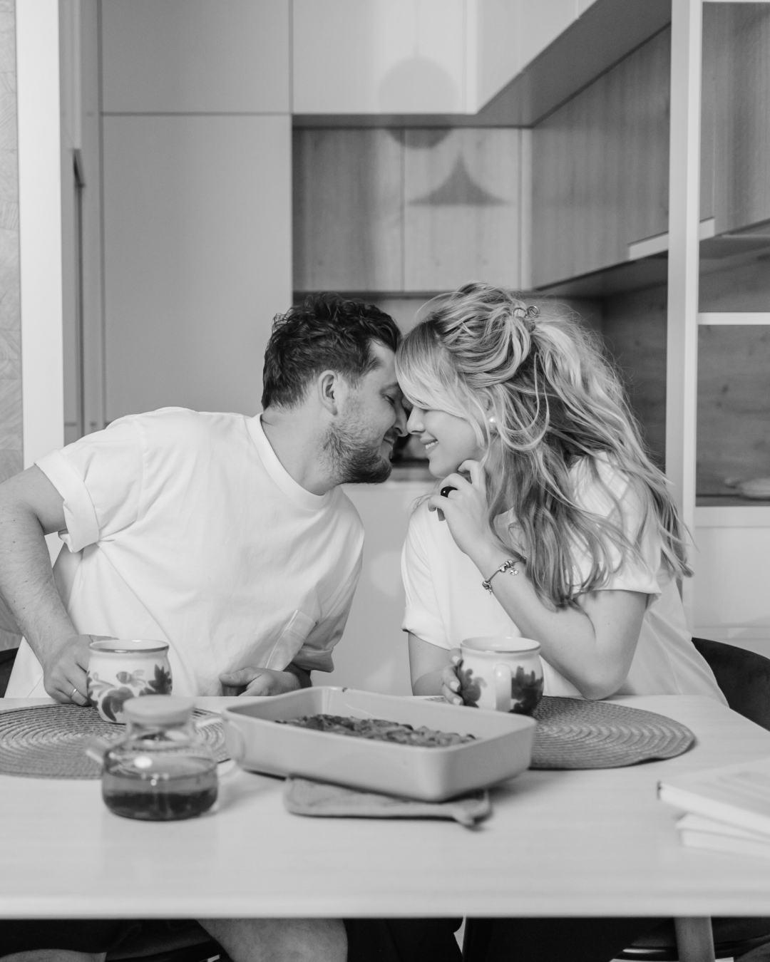 A black and white photo of a couple sharing a tender moment at a kitchen table, their foreheads touching, smiling, with mugs and a baking dish in front of them.