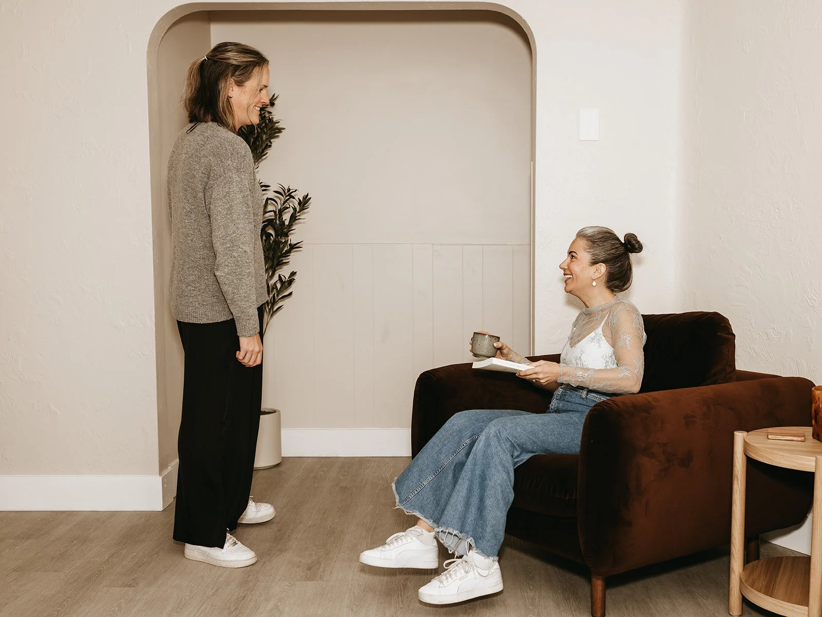 Two women in a cozy living room, one standing and one sitting on a couch, sharing a happy moment. The woman on the couch is holding a mug and some papers, wearing a lace top and jeans. The standing woman is smiling and wearing a sweater and black pants.