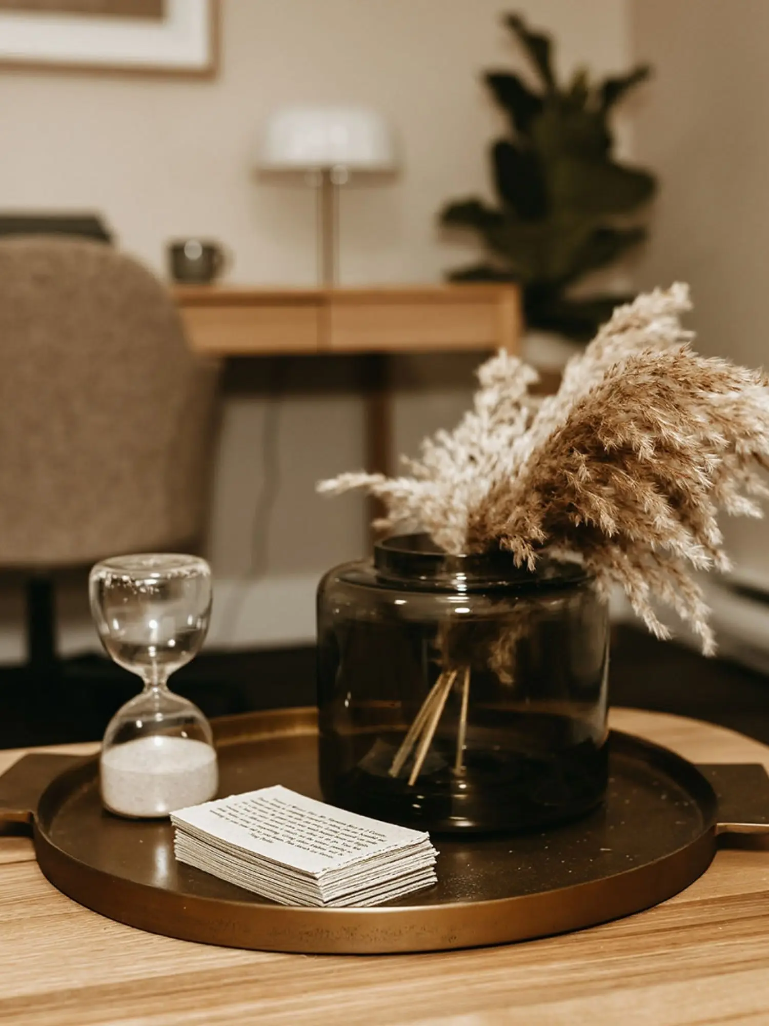 A round tray on a wooden table holding a small hourglass with white sand, a black glass vase with beige dried pampas grass, and a stack of small paper notes. In the background, there's a blurred living room with a beige armchair, a wooden side table, a white table lamp, and a tall potted plant.