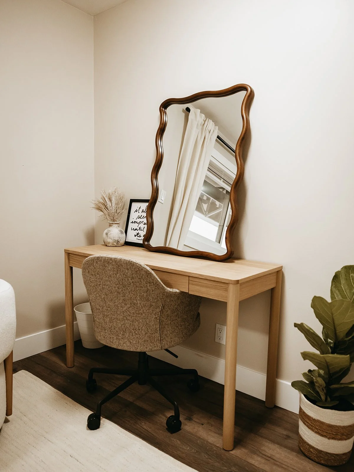 A cozy home office corner with a light wood desk, beige upholstered swivel chair, large wavy-edged mirror, a potted plant, and a framed quote on the desk.