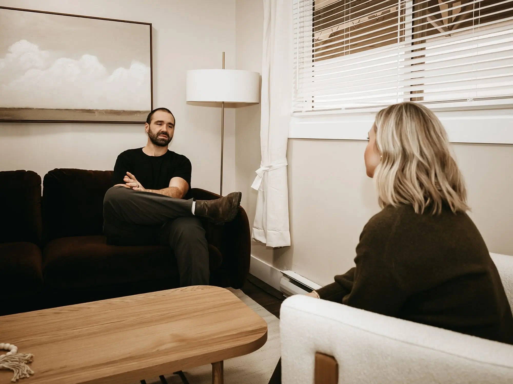 A man and woman having a conversation in a living room. The man is sitting on a dark brown sofa, wearing a black t-shirt and dark pants. The woman is sitting on a light-colored armchair, facing away from the camera. The room has white walls, a window with blinds, and a wooden coffee table.