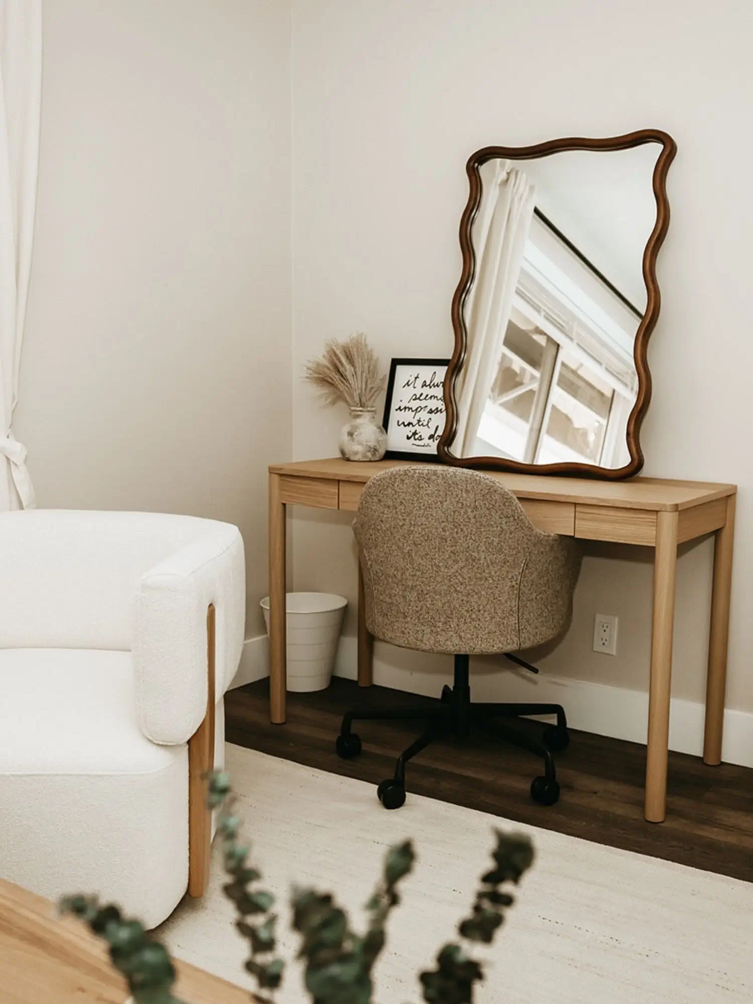 A cozy workspace with a wooden desk, a beige office chair, a mirror, a framed quote, a vase with dried pampas grass, and a small white trash bin. The desk is against a white wall, and there is a window with white curtains reflected in the mirror.