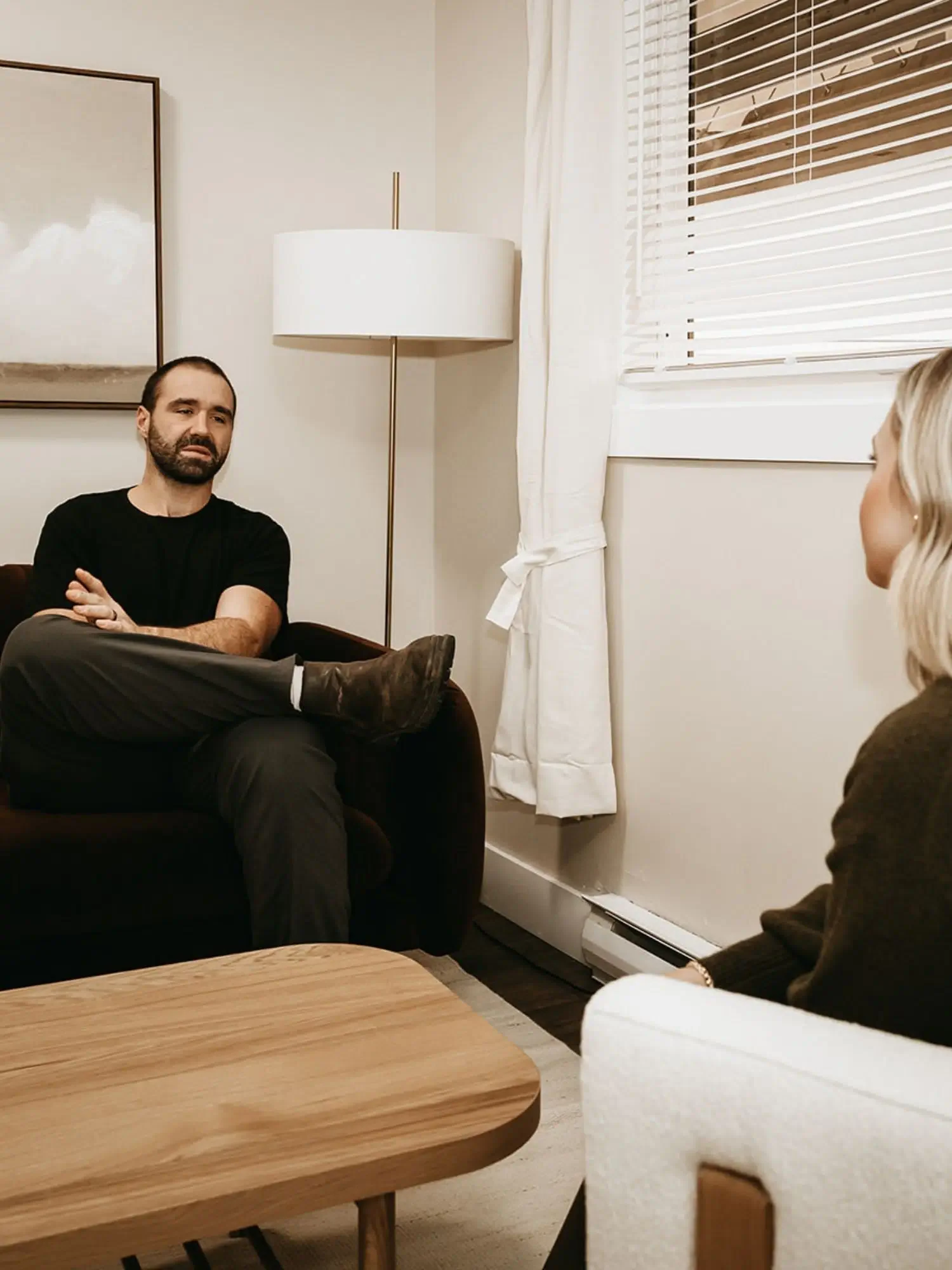 Man sitting with arms crossed on a sofa, talking to a woman in a living room with a window, curtains, a lamp, and wall art.