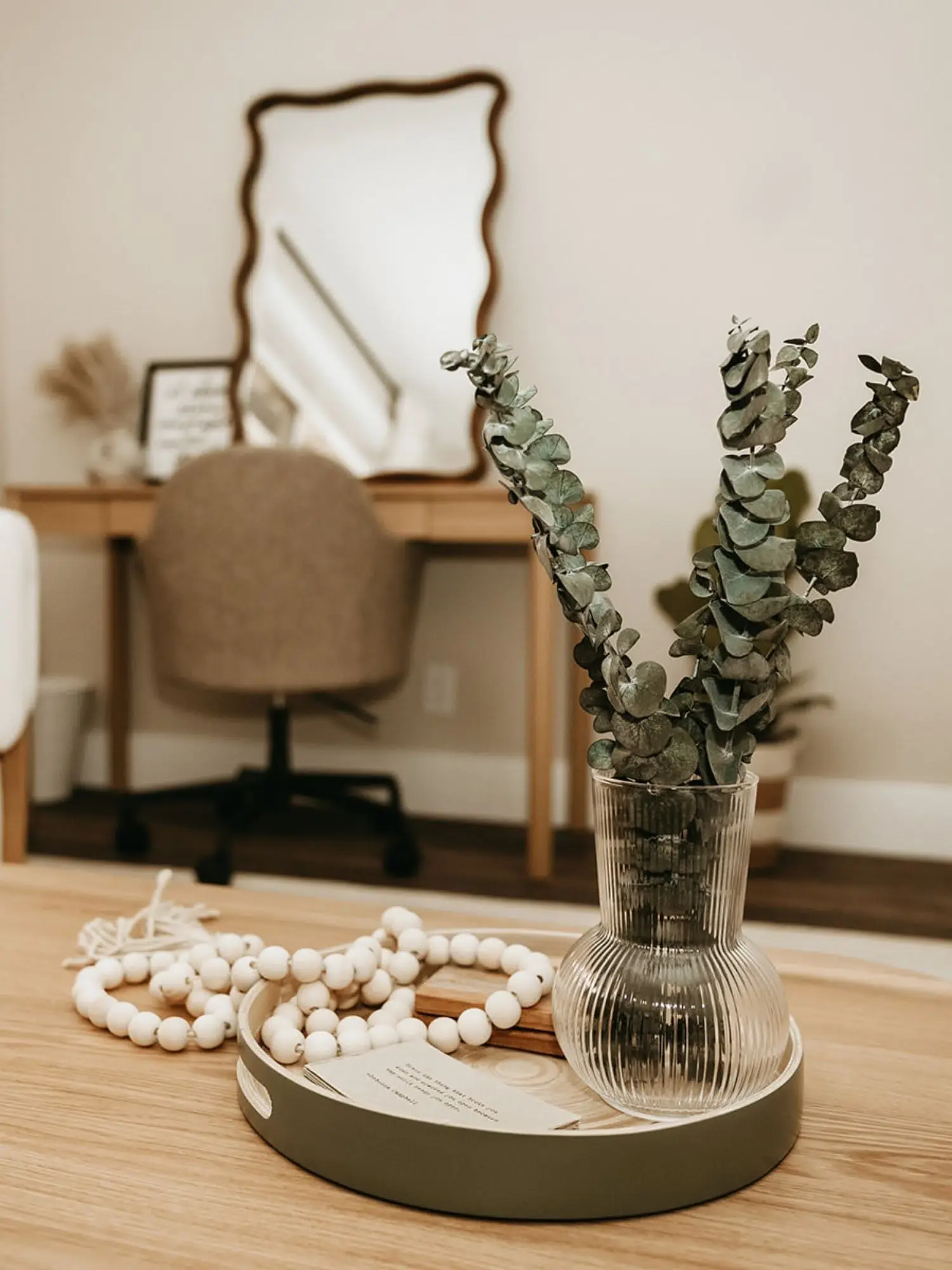Close-up of a wooden table with a clear ribbed glass vase containing eucalyptus branches, a gray tray with a string of white beads, and a small card, with a blurred background of a beige office chair, a wooden desk, and a decorative mirror on the wall.