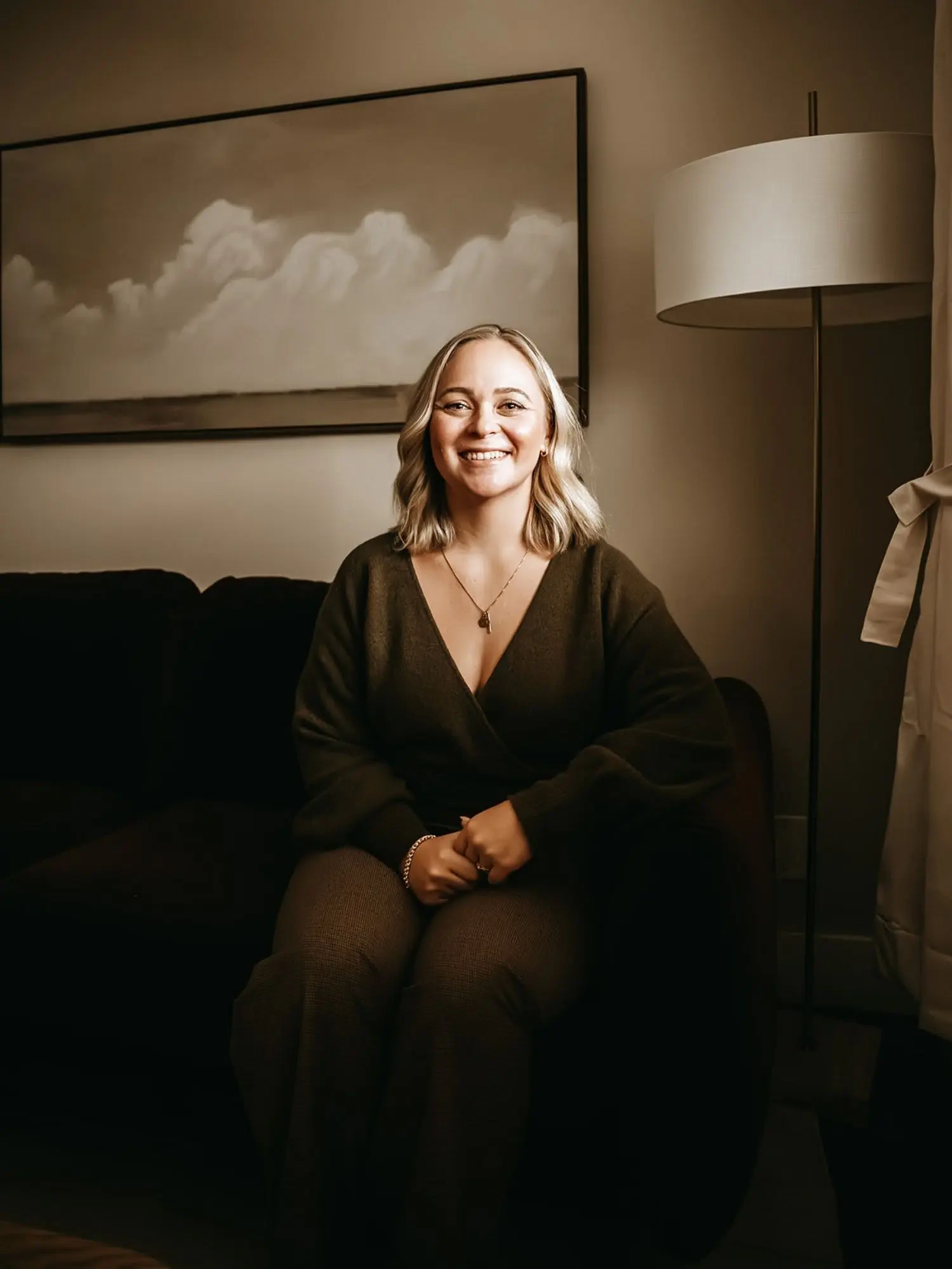 A woman with blonde hair sitting on a black couch, smiling, indoors, with a large framed cloud photo on the wall behind her and a tall floor lamp to her right.