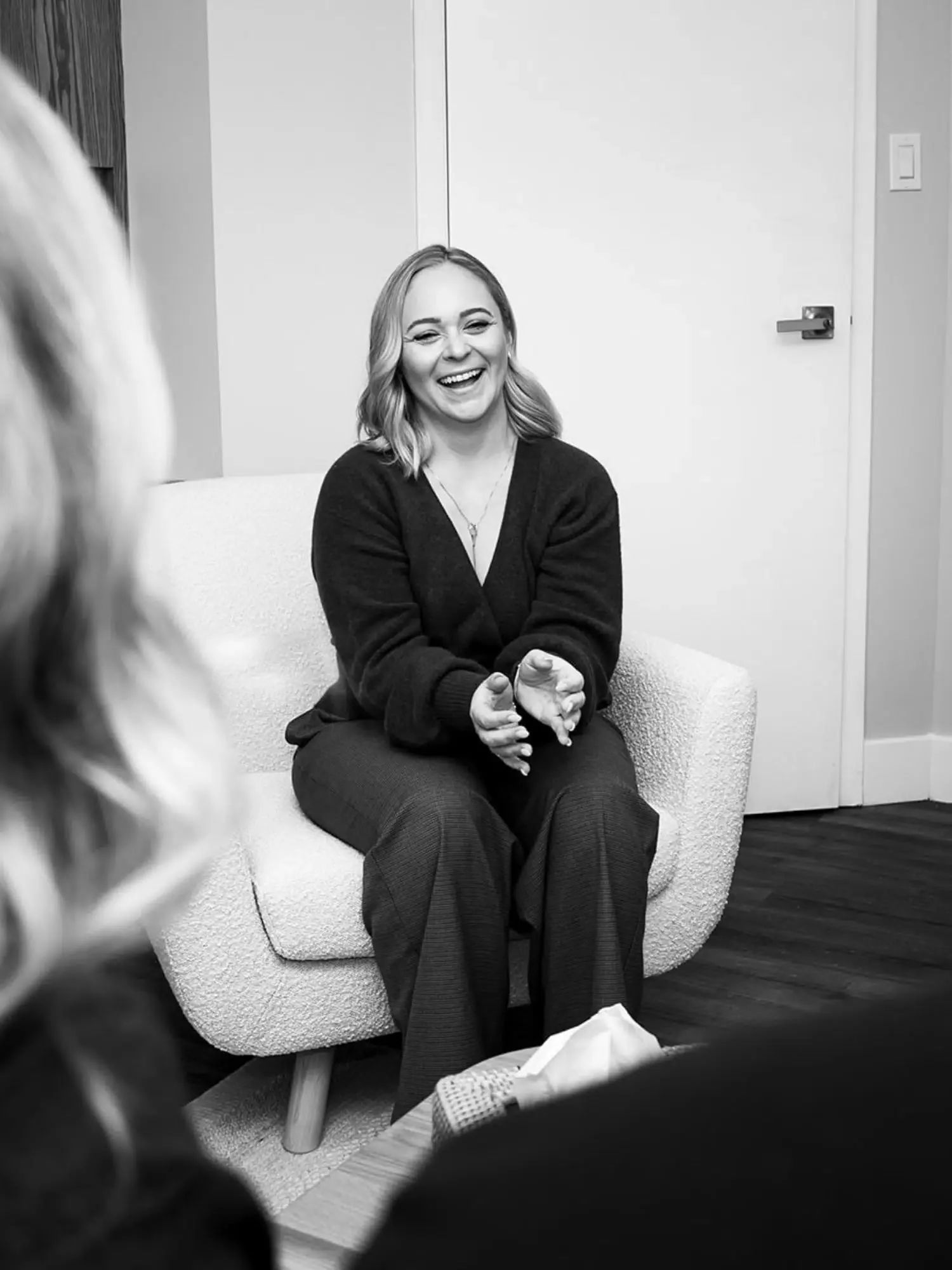 A woman with shoulder-length hair smiling and clapping, sitting on a textured armchair in a room with a closed door in the background, black and white photograph.
