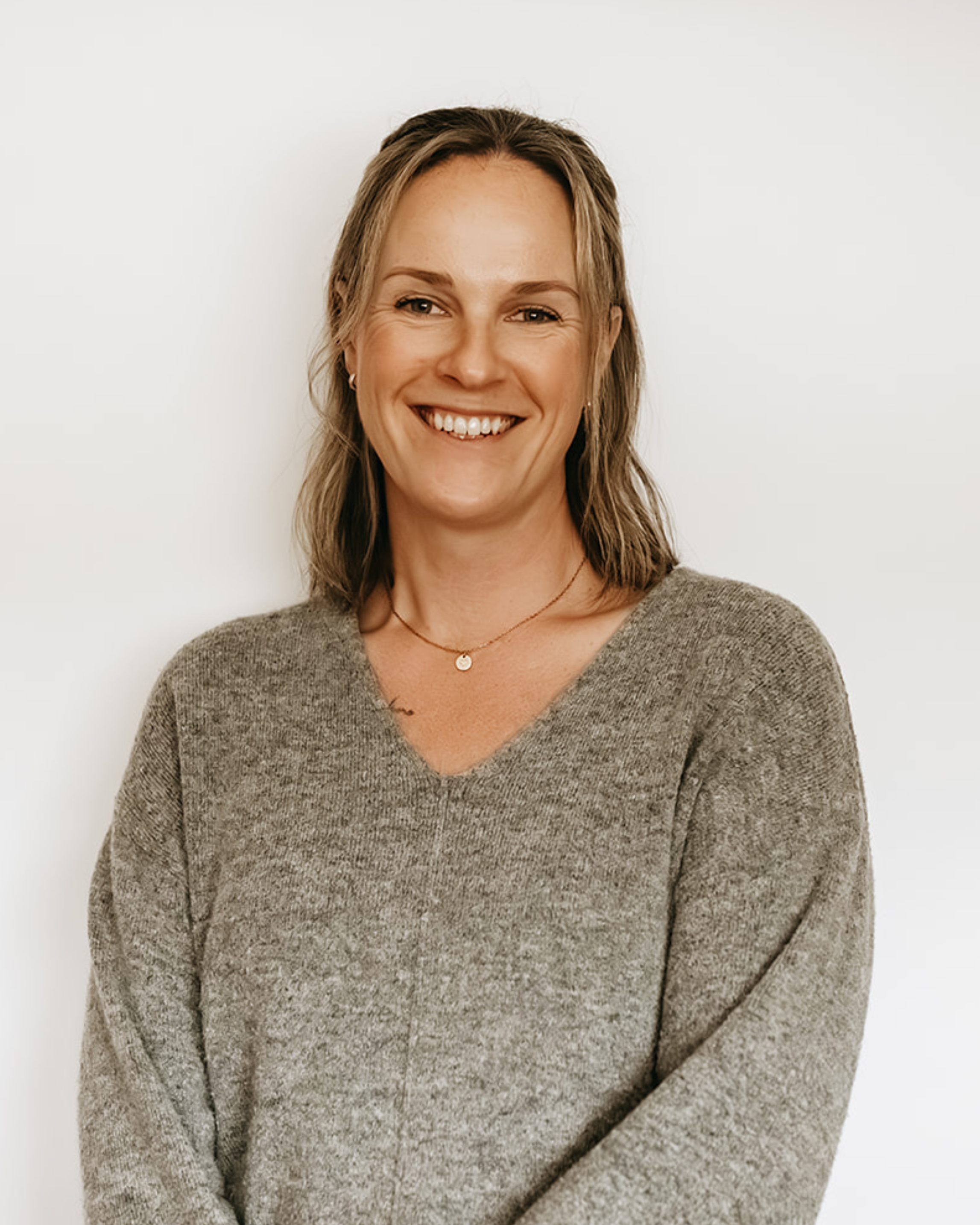 A smiling woman with shoulder-length hair wearing a gray sweater and a delicate necklace, standing against a white background.