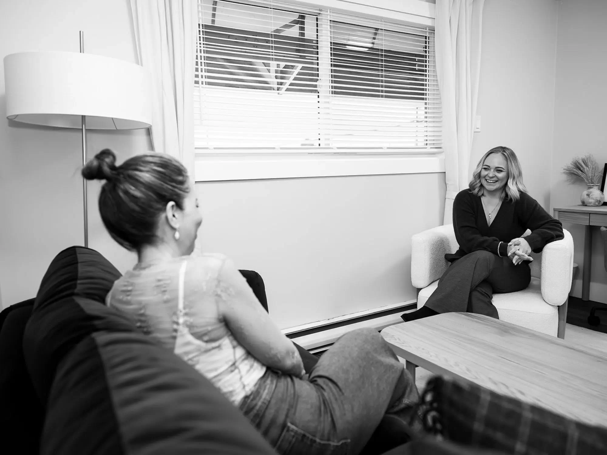 Two women having a conversation in a cozy living room with a window, curtain, and a side table with decorative items.