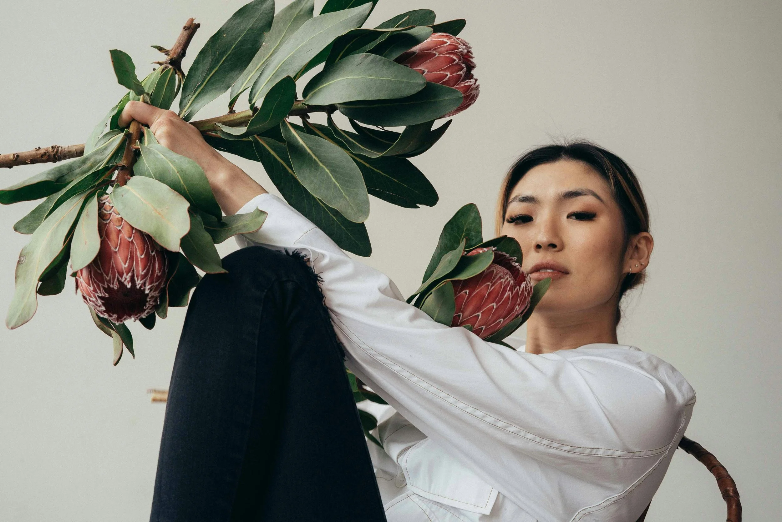A woman with short dark hair and earrings, wearing a white shirt, is sitting against a plain gray background, holding large pink protea flowers and green leaves.