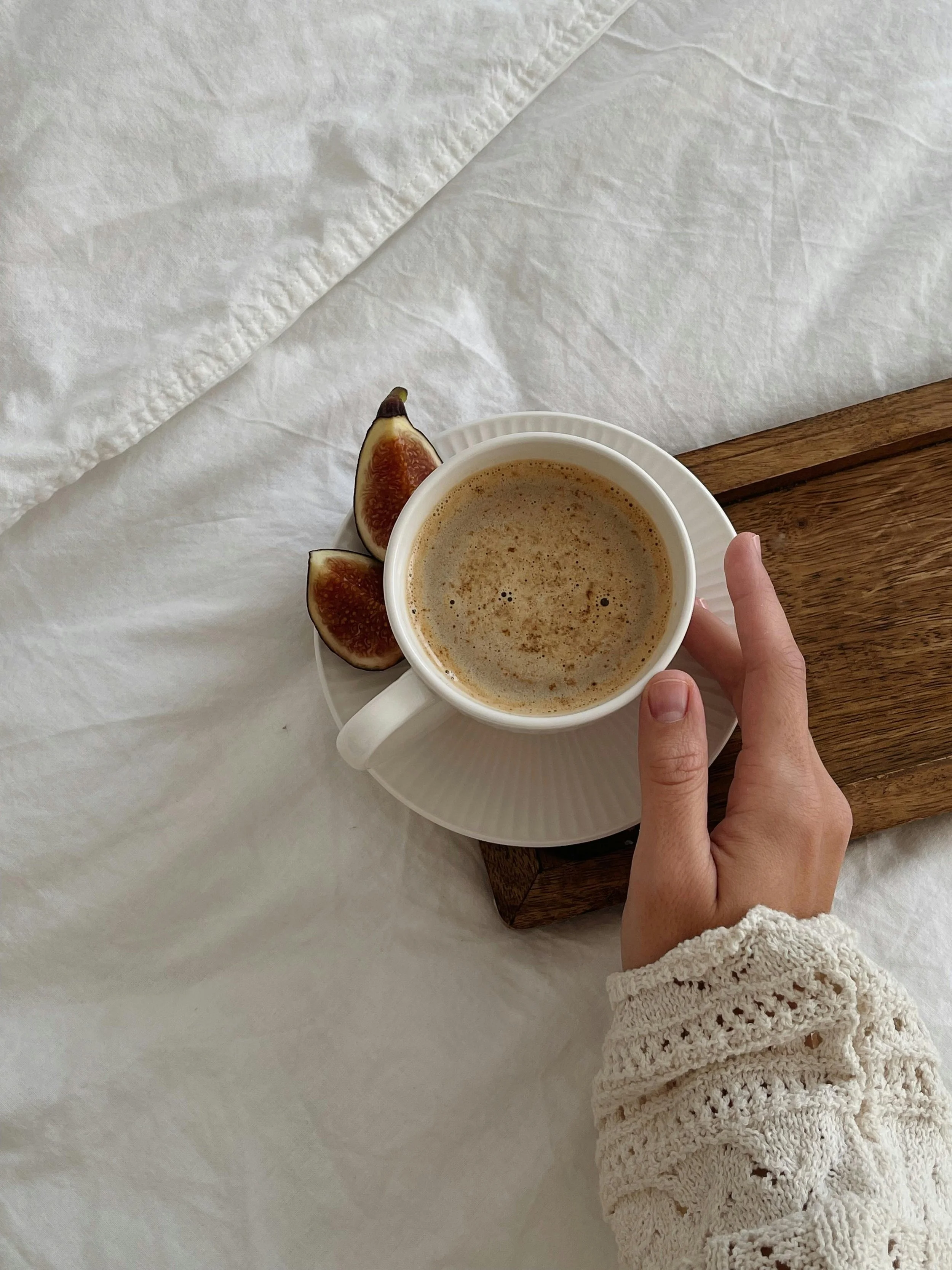A hand holding a white cup of coffee with foam, placed on a matching saucer with two fig halves, all on a wooden tray.