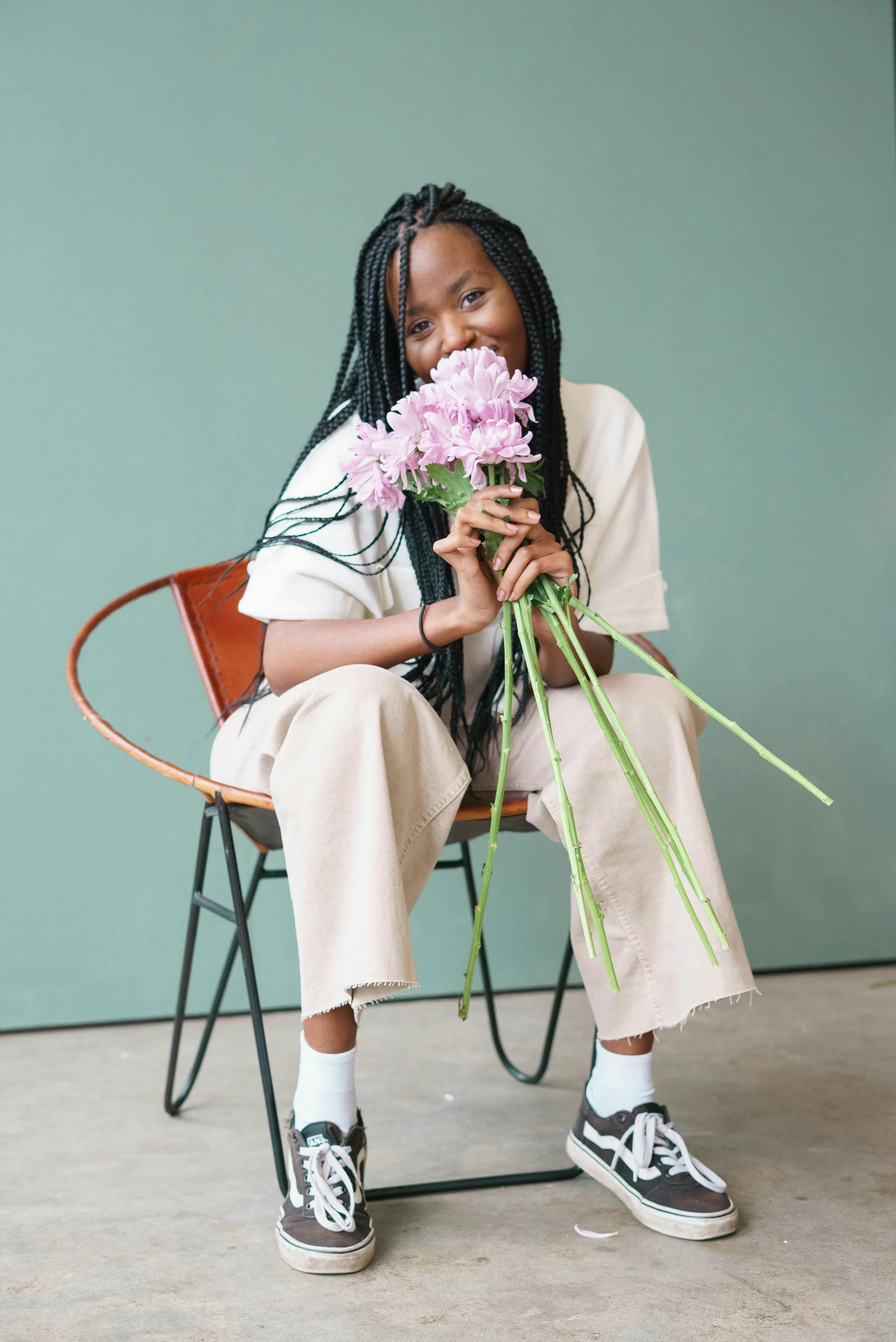 A smiling girl with long black braided hair sits on a chair, holding a bouquet of pink flowers, against a plain green background.