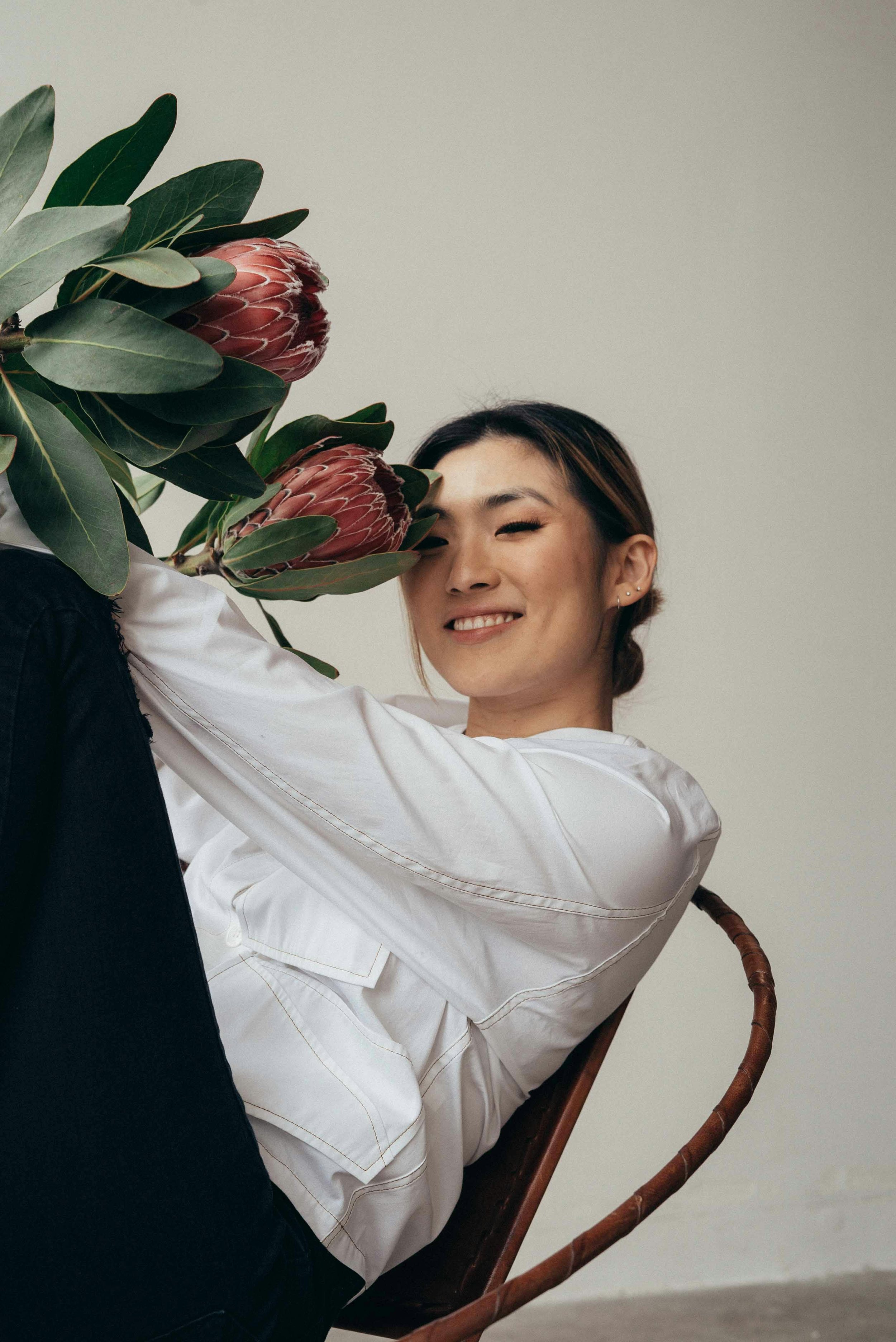 A woman with short dark hair, wearing a white shirt, sitting on a chair, smiling, and holding a large plant with dark green leaves and pinkish flowers close to her face.