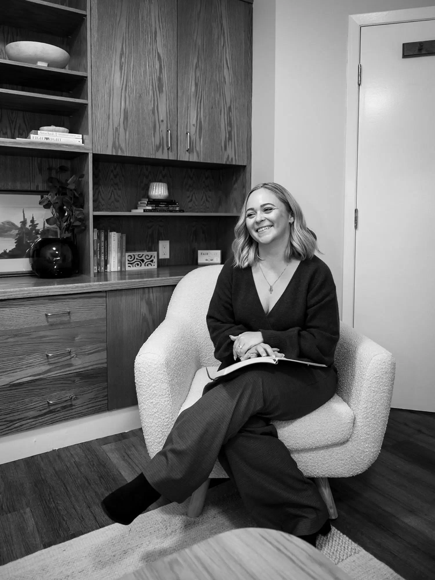 A woman smiling sitting in a cozy armchair with a notebook on her lap in a well-decorated room with books, art, and a potted plant.