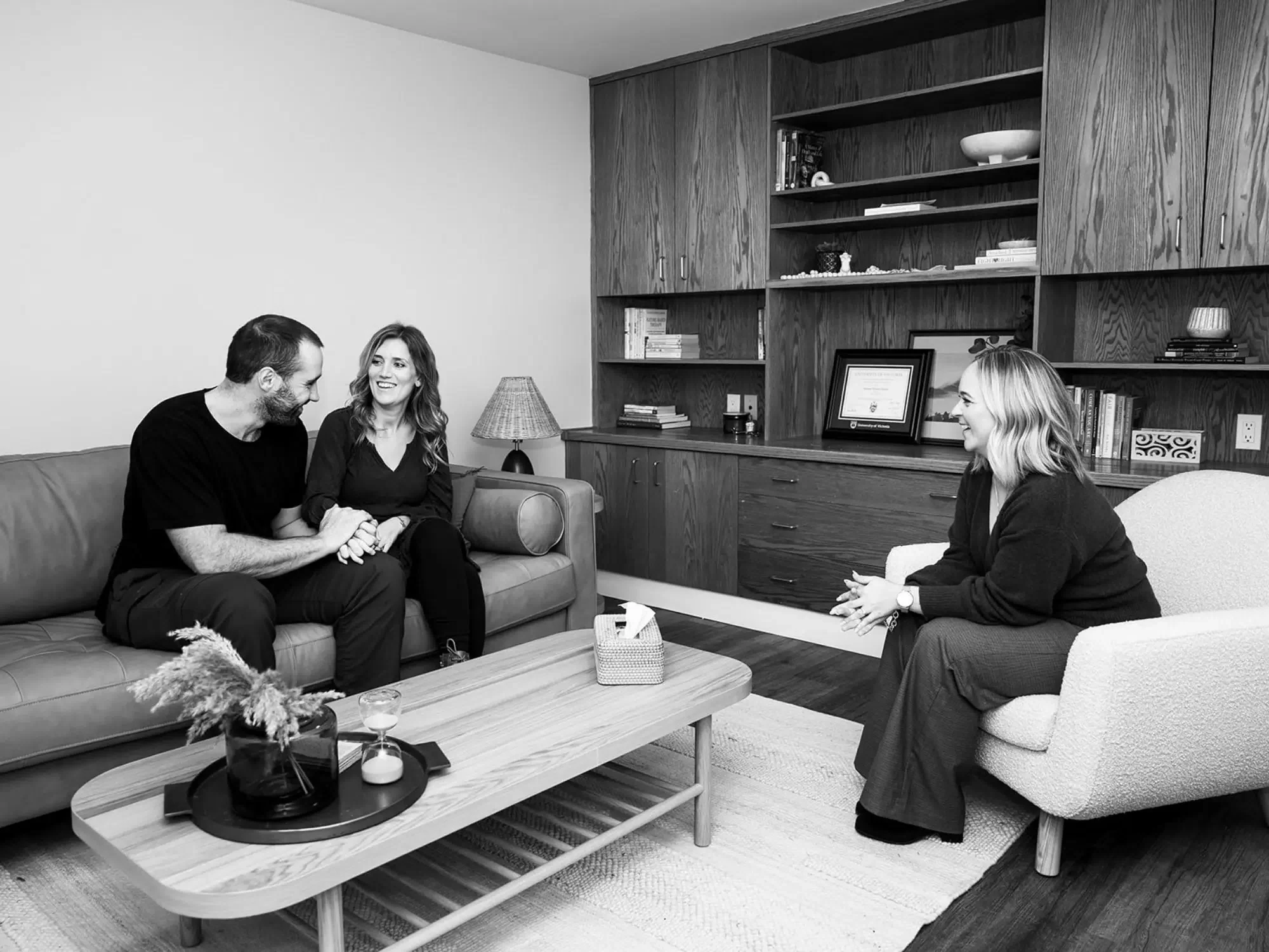 A therapist talking to a couple during a counseling session in a living room with wooden shelves, books, and framed documents.