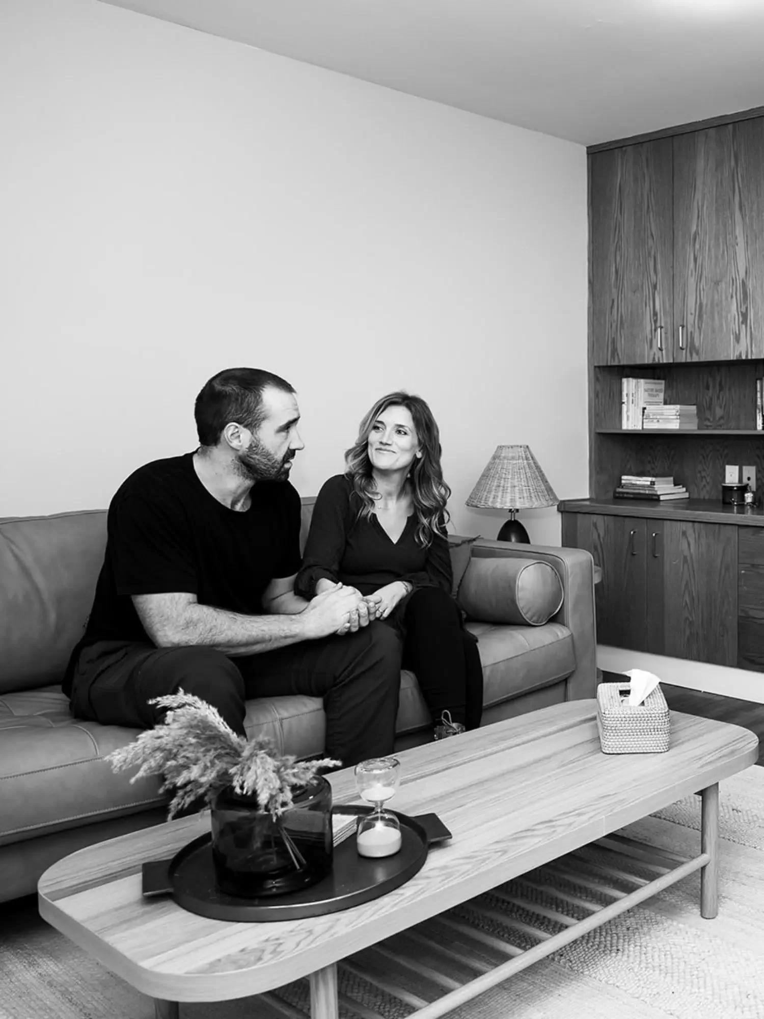 A man and woman holding hands on a sofa in a living room, with a table in front and a bookshelf and lamp in the background.