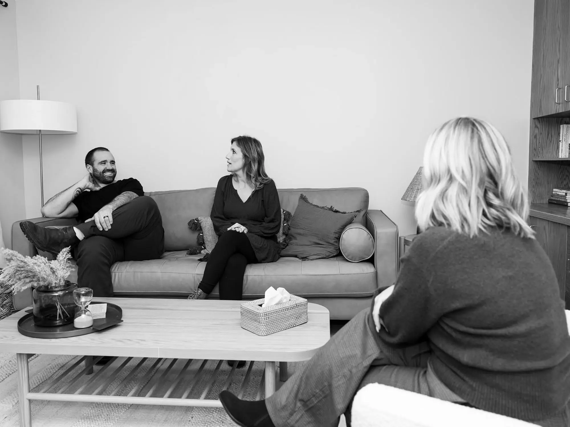 Three women are having a conversation in a living room. One is sitting on a sofa, another on a chair, and the third is partly visible, sitting across from the sofa. The room has a coffee table with a tissue box, a tray with drinks, and decorative items. There are pillows on the sofa, a lamp, and a bookshelf.