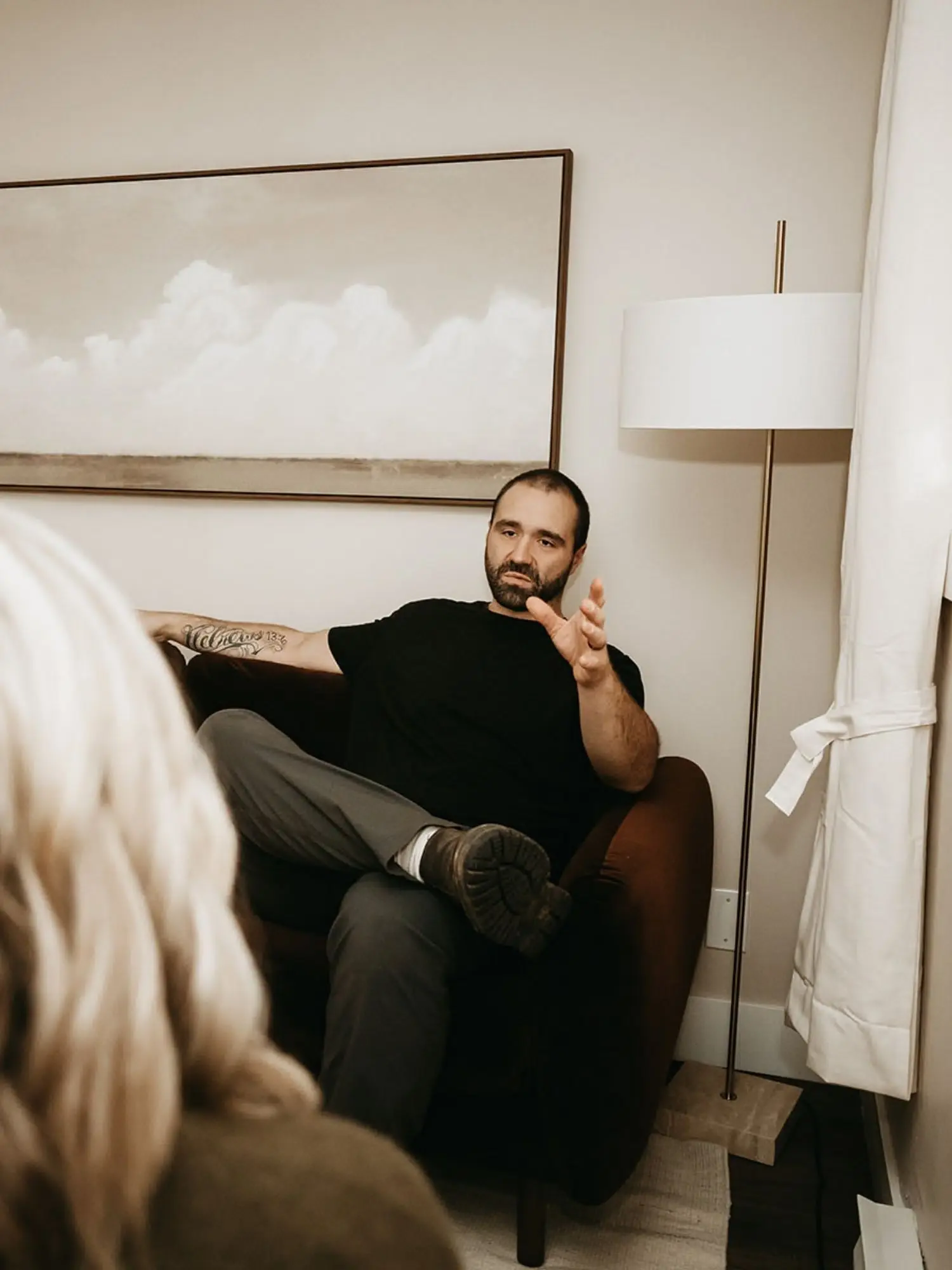 A man with a beard, wearing a black t-shirt and gray pants, sits on a dark-colored armchair in a room with cream-colored walls, a large cloud-themed painting, a white floor lamp, and partially visible curtains, engaging in a conversation.