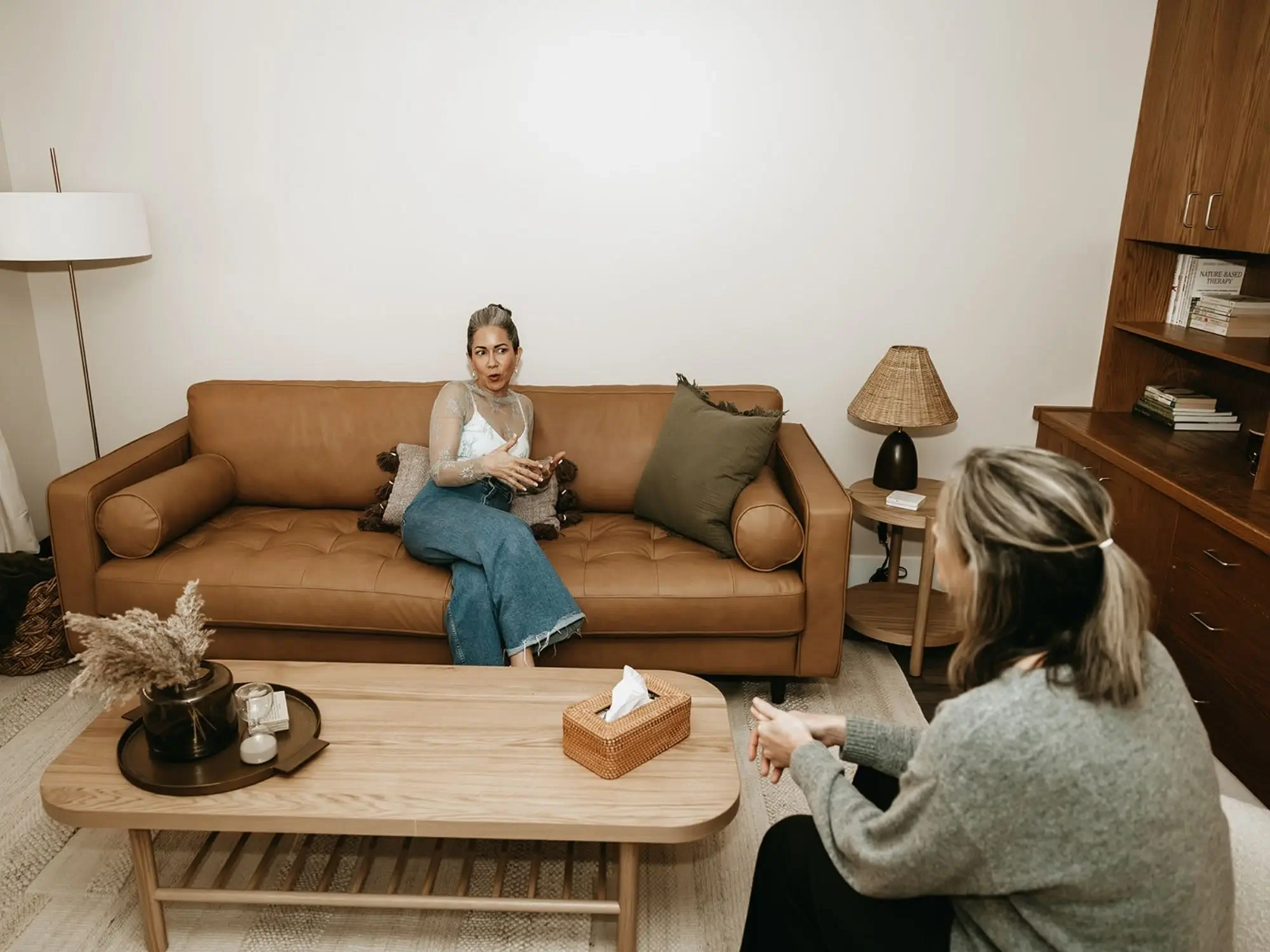 Two women having a conversation in a living room with a brown sofa, coffee table, and bookshelf.