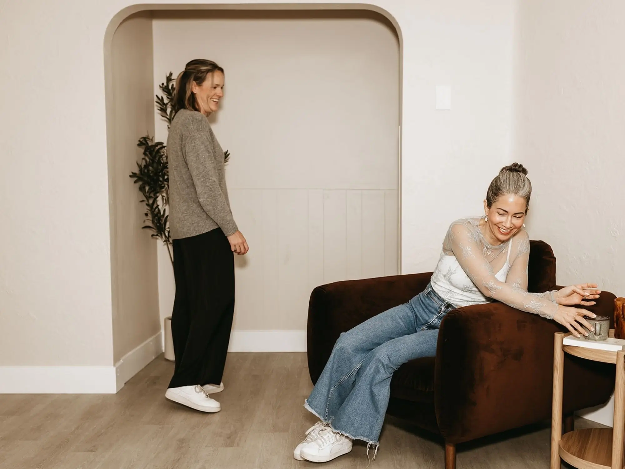 Two women are smiling and laughing in a cozy, well-lit room. One woman is sitting on a brown velvet couch, wearing a sheer, lace top and jeans, with her hair in a bun. The other woman is standing nearby, wearing a gray sweater, black pants, and white sneakers, with her hair down. There is a small wooden side table with a mug and a decorative item on it.