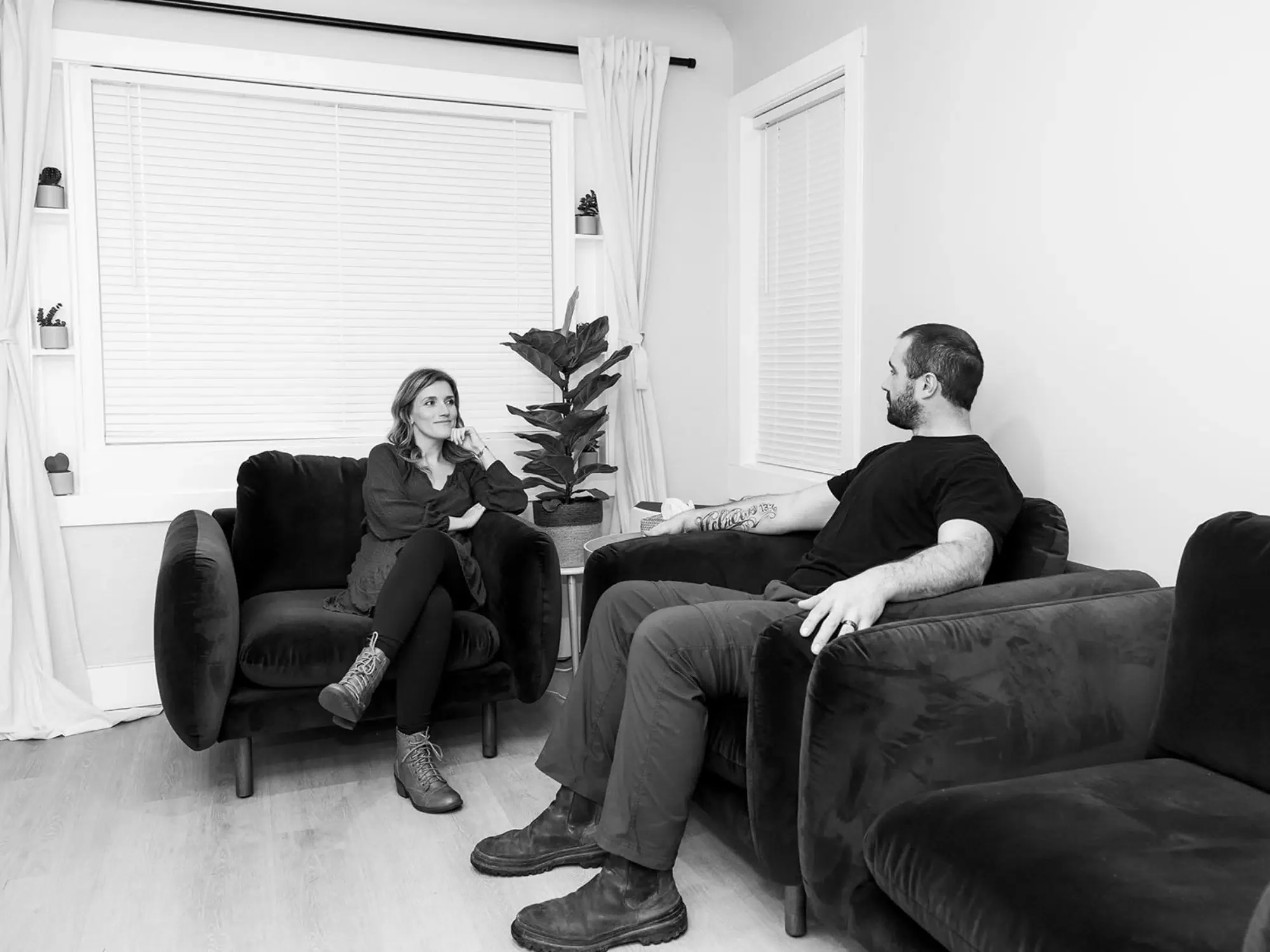Black and white photo of a woman and man sitting on separate couches in a living room, facing each other, engaged in conversation, with a large plant and window behind them.