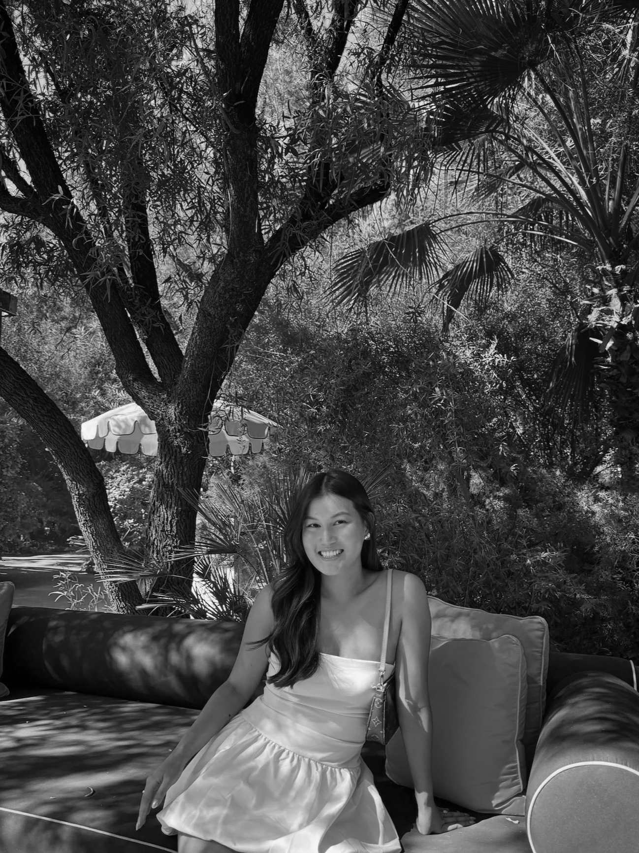 A woman sitting on outdoor furniture, smiling, with trees and a patio umbrella in the background.