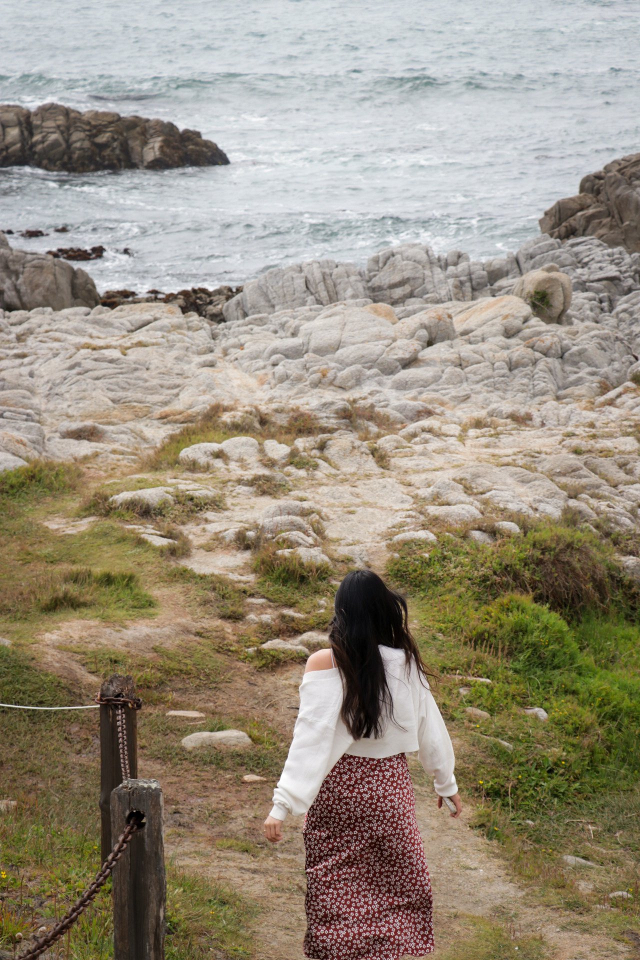 A woman with long dark hair walking along a dirt path through grassy terrain toward rocky cliffs and the ocean.