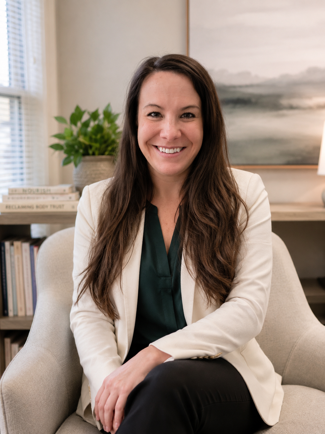 A woman with long brown hair smiling, sitting on a beige armchair in a well-lit room with a bookshelf, potted plant, and abstract painting in the background.