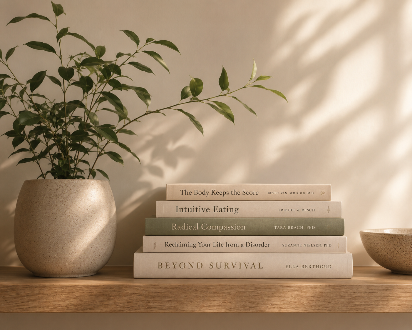 A potted plant with green leaves on a wooden surface next to a neatly stacked collection of six books with beige and green covers. The titles include 'The Body Keeps the Score,' 'Intuitive Eating,' 'Radical Compassion,' 'Reclaiming Your Life from a Disorder,' and 'Beyond Survival.'